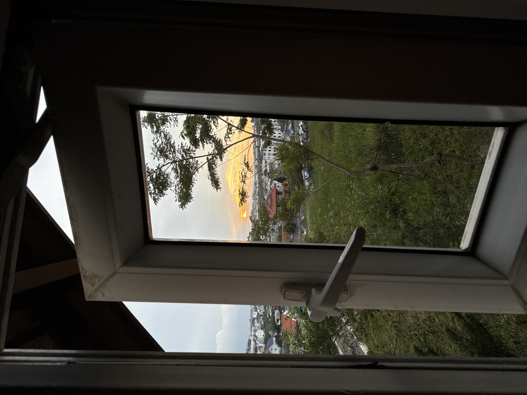 The image captures a view of Lâm Viên - Đà Lạt, Vietnam, through an open white-framed window. The window handle is visible. The scene outside is split by the horizon, with a sunset visible above buildings and trees. Below the horizon, a residential area with houses is visible. The foreground shows the window frame and portions of the interior. The overall lighting is dim inside, contrasting with the bright sky outside.