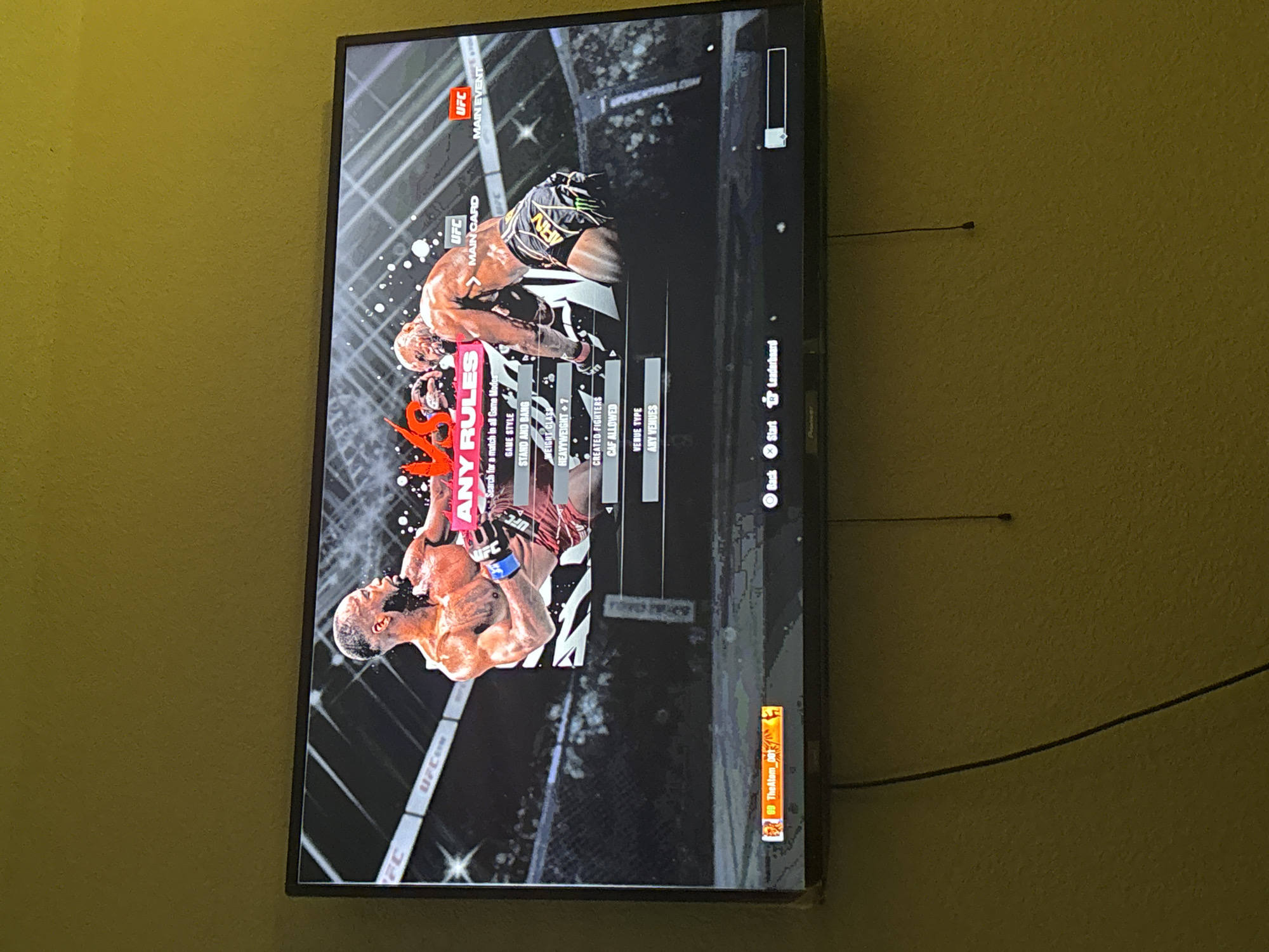 A flat-screen television displaying a video game is mounted on a wall. The game appears to be a UFC (Ultimate Fighting Championship) fighting game, as evidenced by the UFC logos present on the screen. The menu displayed shows options for game style, weight class, whether created fighters are allowed, and venue type. Two fighters are prominently displayed in the center of the screen in an action pose, surrounded by a blurred, stylized background resembling a fight arena. The text "ANY RULES" is overlayed in a graphic style, implying a game mode with minimal rule restrictions. UI elements such as "Back," "Start," and "Leaderboard" are located on the right side of the screen. The screen displays the player name "TheAtom_DOT." The wall is painted a solid yellowish-green color. An antenna is visible behind the TV. The TV is located in an unspecified location within Arlington, United States of America.