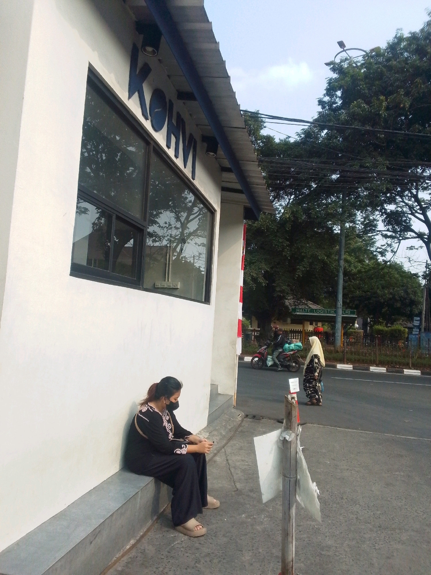A woman wearing a black long-sleeved shirt, black pants, and tan slip-on sandals sits on a concrete ledge outside a white building with a dark blue sign that reads "KOHVM". She is wearing a black face mask and looking down at her phone. In the background, a person wearing a yellow hijab and a patterned dress stands on the side of a road, and a motorcycle with a driver wearing a black jacket and helmet drives past. A traffic light pole and trees are visible. The scene appears to be in a public area, possibly near a business or logistics center, as suggested by a partially visible sign that reads "HALTE LOGISTIKOLA". The overall lighting indicates it is daytime.