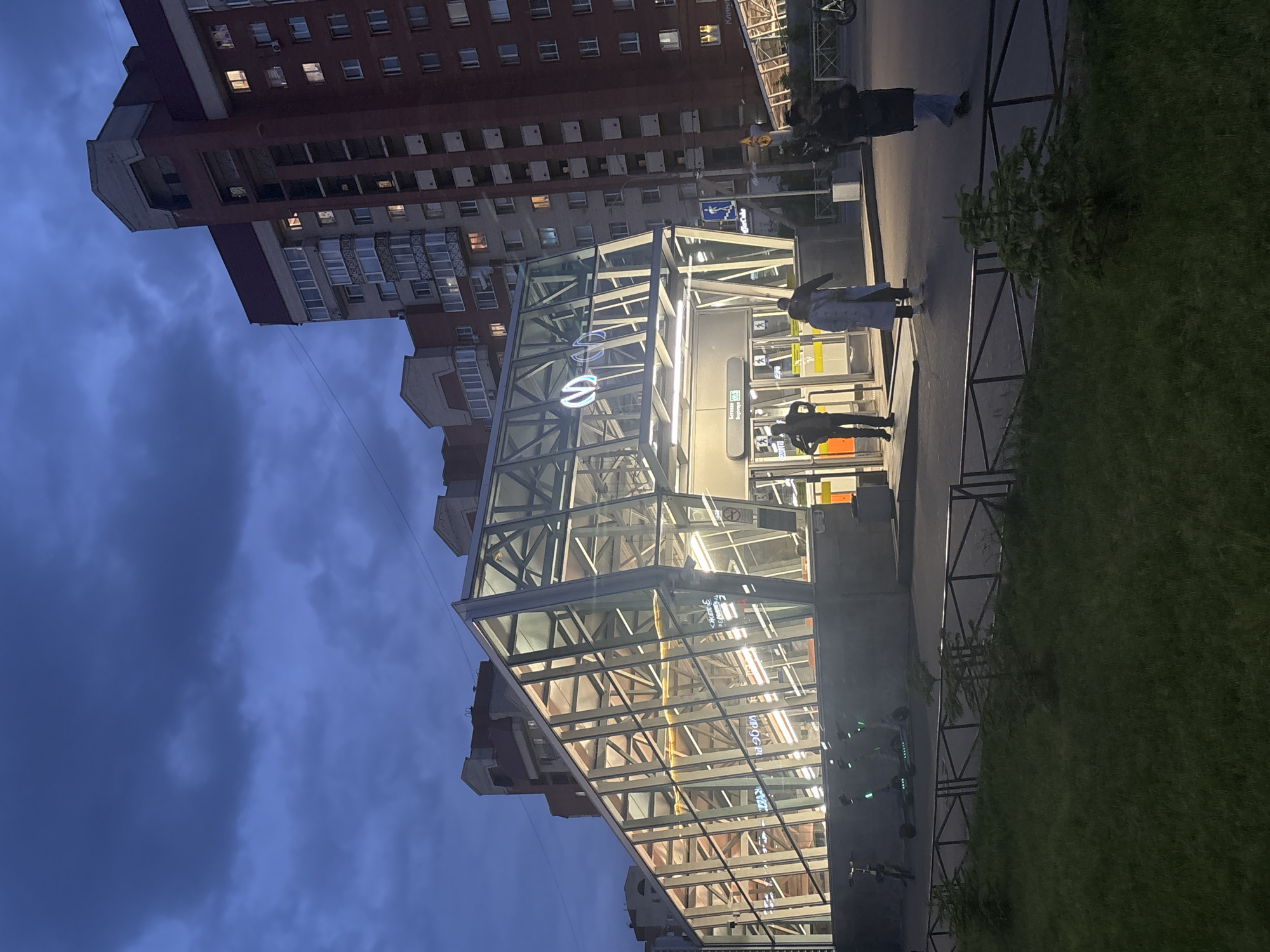 The image shows the entrance pavilion of the Begovaya metro station in Saint Petersburg, Russia, at dusk. The glass and metal structure of the pavilion is brightly lit, contrasting with the darkening sky. A tall apartment building is visible in the background. Several people are walking near the entrance. A row of electric scooters is parked along the sidewalk. A low metal fence separates the walkway from a patch of grass.