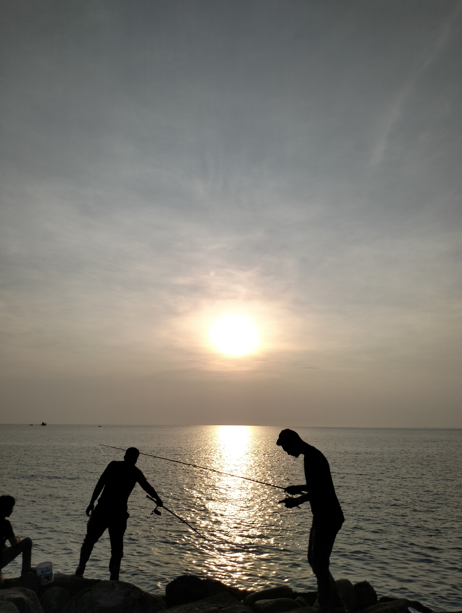 Two people are silhouetted against a sunset over the ocean. They are fishing, with fishing rods extended over the water. A third person is seated, possibly observing. The location appears to be a rocky shoreline near the sea. A small boat is visible in the far distance. The image is captured in the vicinity of Lhokseumawe, Indonesia.