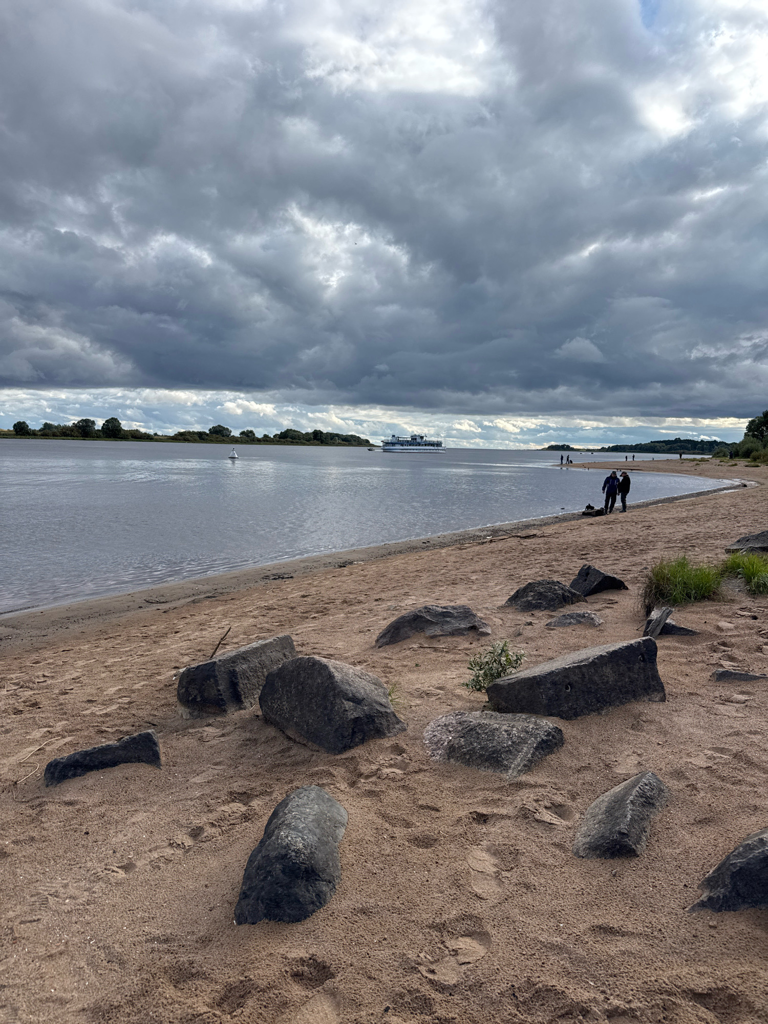 The image depicts a cloudy day at a sandy beach on the Volkhov River in Veliky Novgorod, Russia. The foreground is dominated by scattered dark rocks and sand, with faint footprints visible. In the middle ground, the calm river stretches towards the horizon, bordered by a distant treeline. A white buoy marks the water. Further out, a white passenger ship is visible, along with several figures walking or standing on the sandy shore of the far bank. Two individuals are standing close together in the middle ground on the sandy beach. The sky is heavily overcast with dark, dramatic clouds.