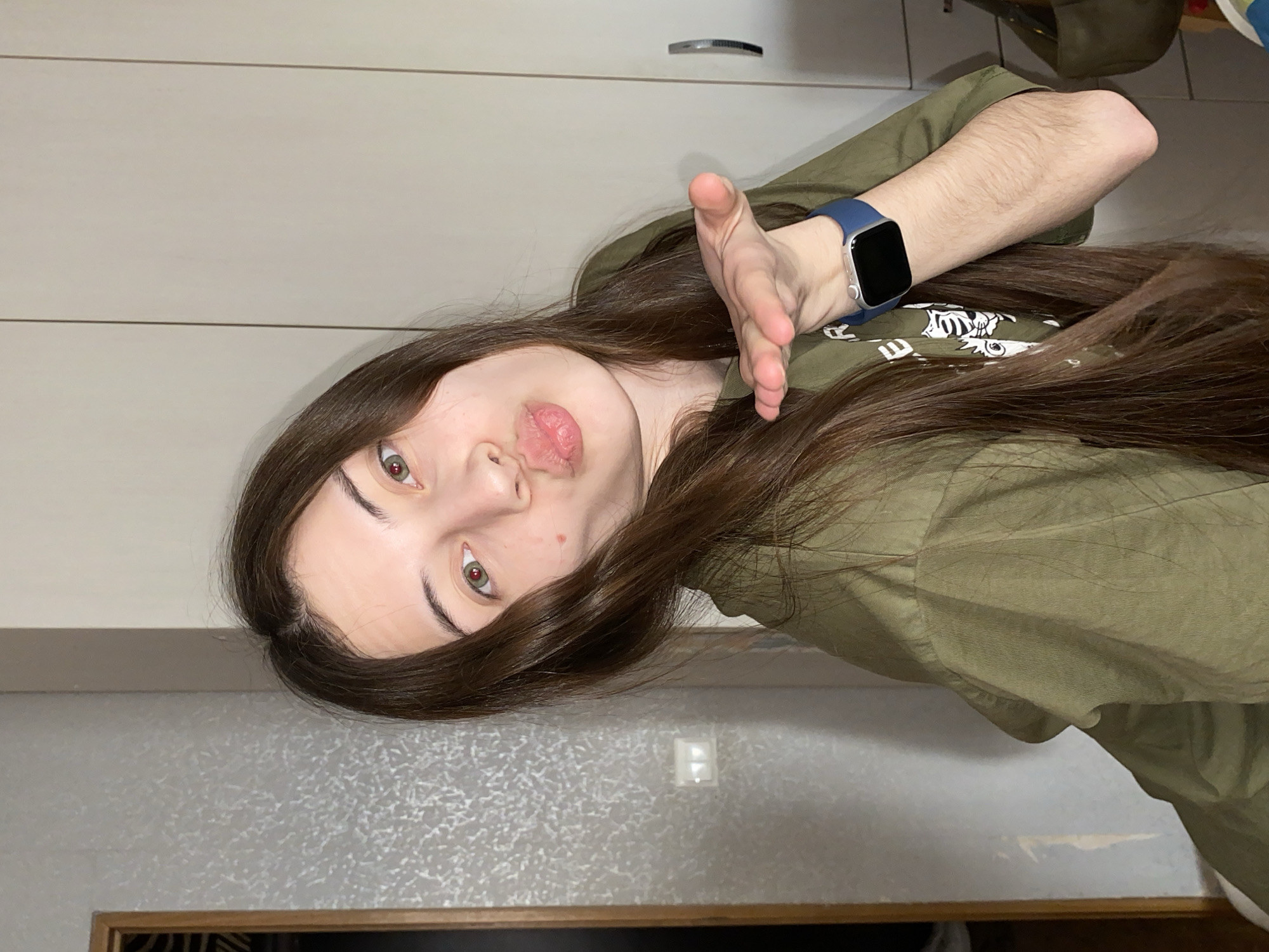 This media file shows a young person indoors in Votkinsk, Russia, captured in a portrait format that is rotated 90 degrees clockwise. The subject has long, dark brown hair that cascades around their face and over their shoulder. They have fair skin, prominent eyebrows, and light-colored eyes that appear hazel or green, exhibiting red reflections, possibly due to flash or direct lighting. Their lips are slightly pursed in what could be interpreted as a 'kiss face' or playful expression.
They are wearing a muted olive green long-sleeved top, and a blue smartwatch with a silver frame and black screen is prominently visible on their left wrist. Their left arm is extended towards the camera, with the palm open in a gesturing motion. The forearm also shows noticeable hair.
The background suggests a home environment. To the left of the subject's head (which would be above them if the image were upright), there is a light beige wall with horizontal paneling or cabinet lines and a silver drawer handle. To the right (below the subject), there is a greyish wall with a textured, speckled pattern and a white light switch. A wooden frame or edge is visible at the very bottom of the image. The setting appears to be a casual indoor space. No legible text is visible in the image.