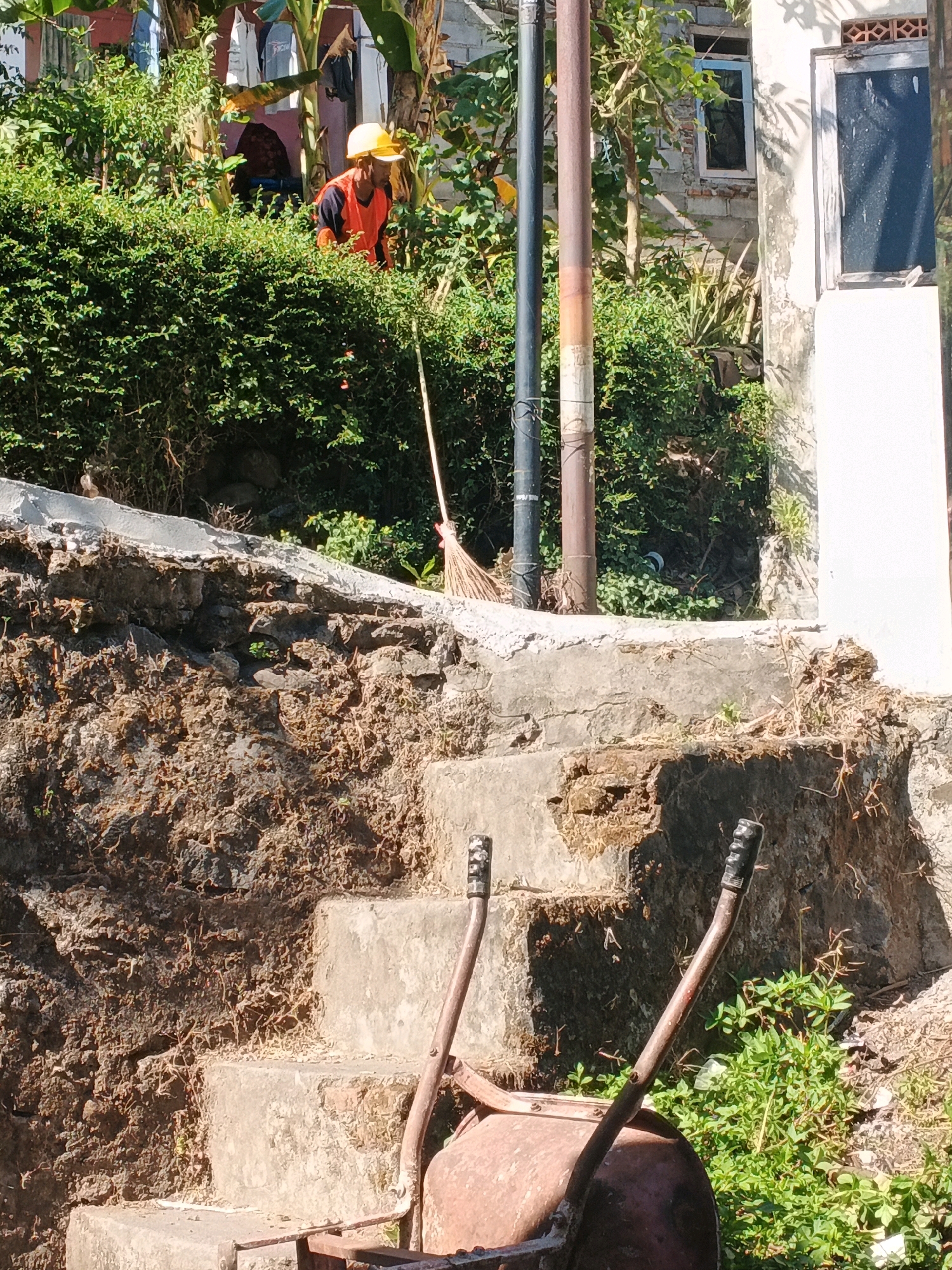 A man in an orange safety vest and yellow hard hat works near a bush-lined retaining wall and concrete stairs in Keji, Indonesia. He appears to be tending to the foliage or performing landscaping. A broom leans against a metal pole behind him. In the foreground, a rusty wheelbarrow sits on the stairs. The scene depicts an outdoor environment with a building visible in the background, bathed in sunlight.