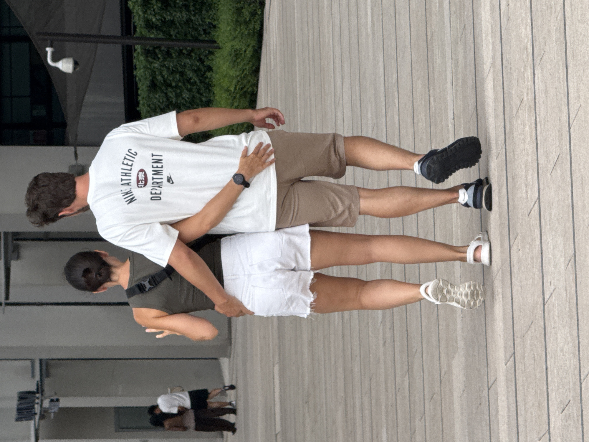 Two individuals, a man and a woman, walk away from the camera on a paved outdoor walkway. The woman, on the left, wears a dark green tank top and white denim shorts. She has her left arm around the man's waist. The man, on the right, wears a white t-shirt with "NIKE ATHLETIC DEPARTMENT" printed on the back, and tan shorts. His right arm is around the woman's waist. The man is wearing a black watch on his left wrist. In the background, to the left, two other people are walking away from the camera. The environment appears to be an urban setting with modern architecture, including a building with grey panels and a security camera visible. The walkway is made of light-colored rectangular paving stones. The location is identified as Yuzhong District, China.