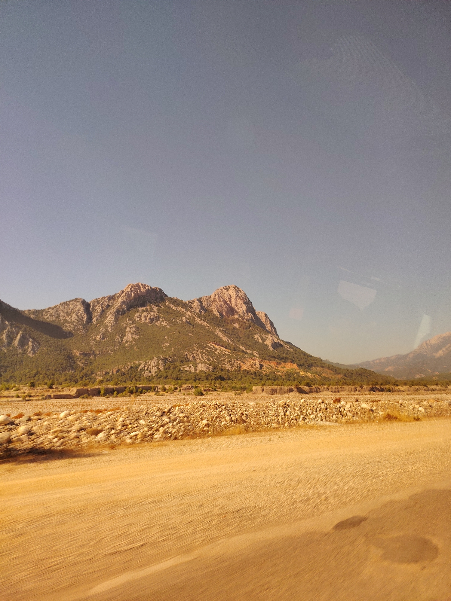 A wide shot captures a mountainous landscape under a clear, pale blue sky. In the foreground, a dry, sandy, and rocky terrain stretches out, appearing to be a riverbed or arid plain. The ground is covered with small stones and pebbles. Beyond this arid area, the middle ground features a low-lying line of sparse vegetation and trees, contrasting with the rocky ground. The background is dominated by rugged mountains, their slopes partially covered with green vegetation and exposed rock formations. The lighting suggests it is daytime, likely late morning or early afternoon, with the sun casting shadows on the mountains. The image has a slightly blurred quality, possibly due to motion or a low-resolution camera. There are no people or animals visible in the scene. The environment appears to be natural and undeveloped. The location is identified as Konyaaltı, Turkey.