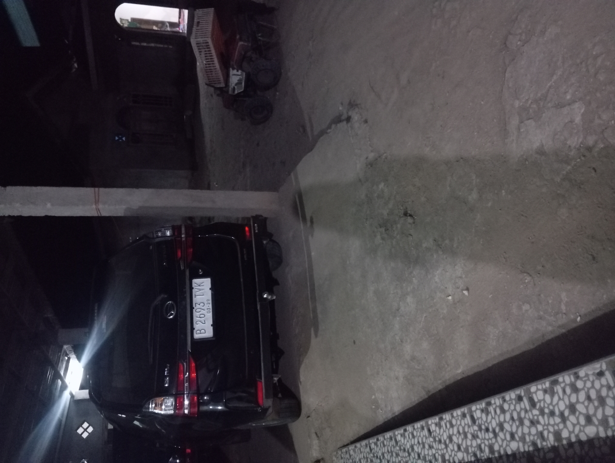 The image shows the rear of a black Daihatsu Xenia car parked on a dirt or concrete ground, likely at night. The car's license plate, B 2693 TYK, is clearly visible. To the right of the car, a small red tractor with a white crate on its back is also parked. The scene appears to be outdoors, possibly in a residential or storage area, with a partially constructed building in the background. The lighting is dim, with some artificial lights casting bright beams. The overall environment suggests a rural or semi-urban setting.