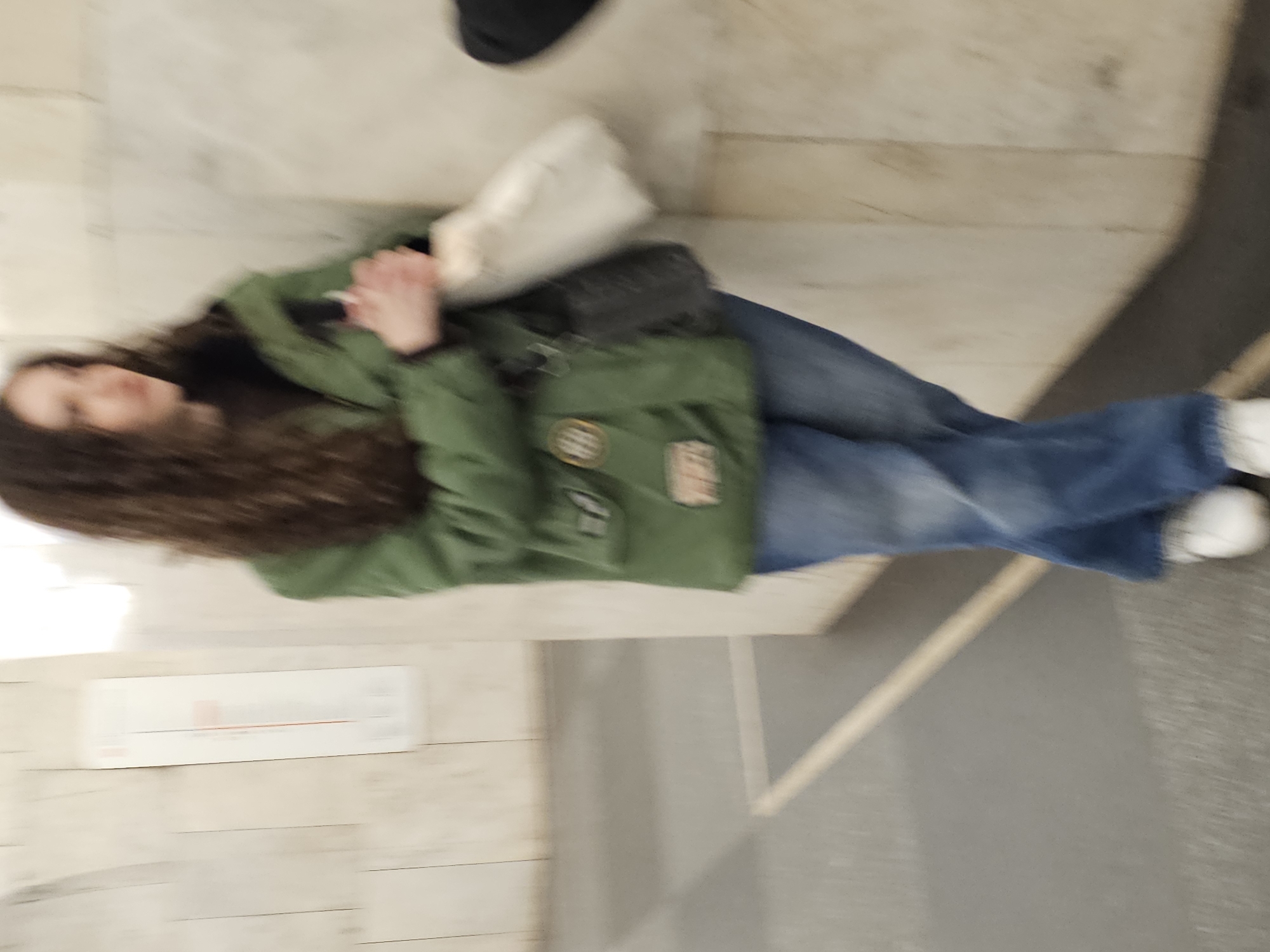 A person with curly brown hair is standing in the Moscow Metro, Russia. They are wearing jeans, white shoes, and a green jacket. They are holding a white shopping bag and a black purse. The subway station has walls made of marble. A railing is visible in the foreground.