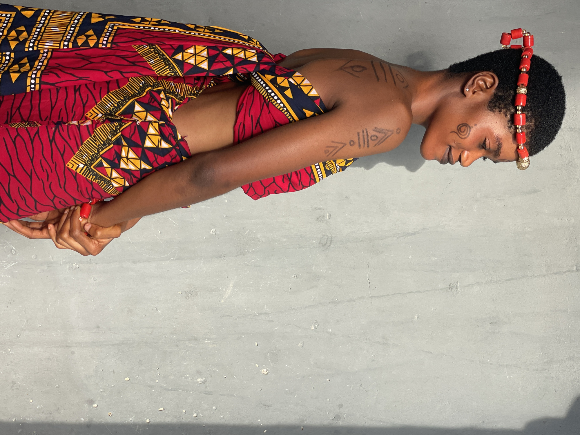 An individual rests in what appears to be a resting or meditative state on a gray surface. The individual has short, dark hair and is adorned with a red beaded headpiece. The person's skin tone is medium brown, and tribal-like markings are present on the face, arm, and shoulder. The individual is wearing a vibrant, patterned fabric, predominantly in shades of red, yellow, and black. The fabric appears to be wrapped around the torso, exposing the midriff. The hands are clasped together.
Without more information, providing a location in Badagry, Nigeria is not possible.