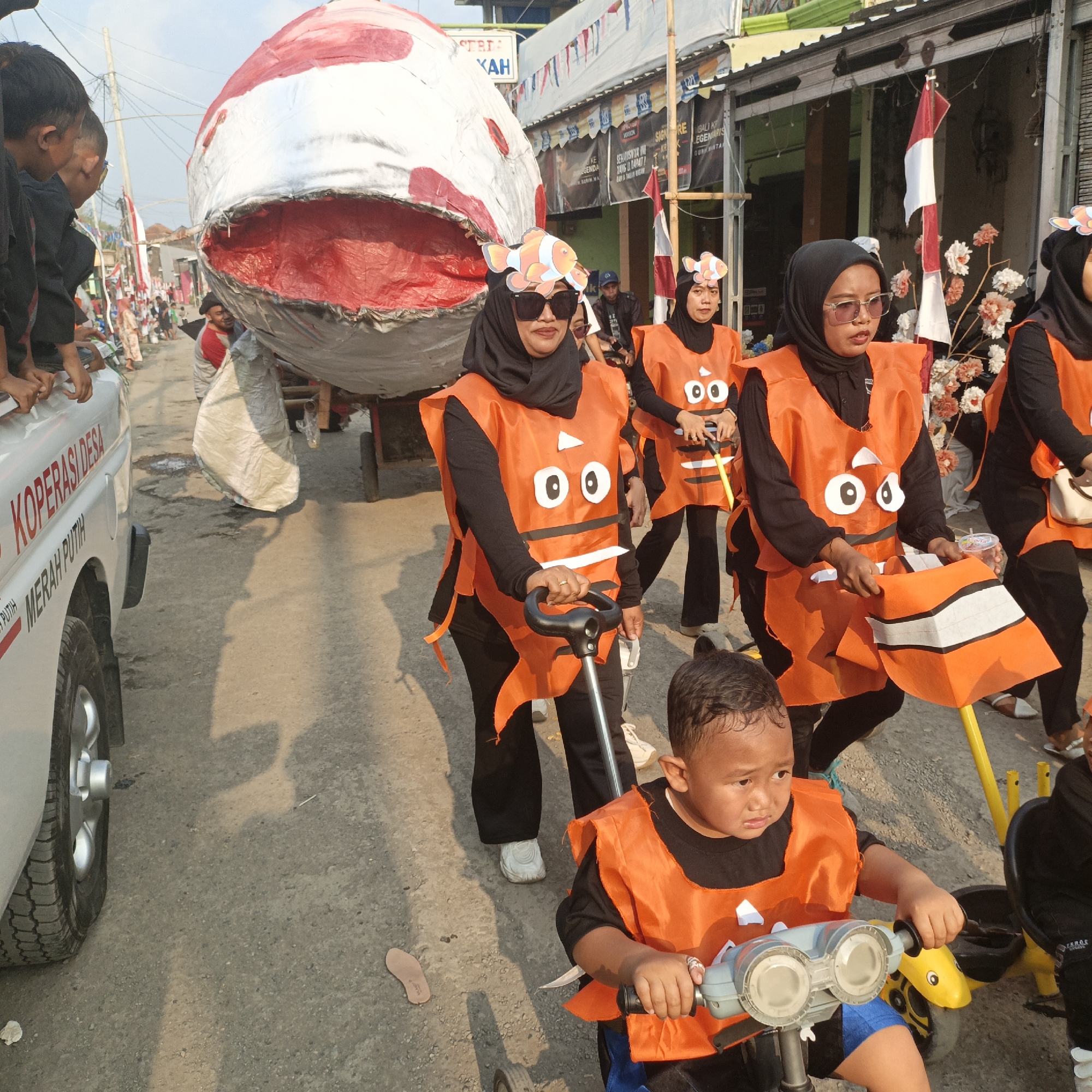 In Tanahbaya, Indonesia, a parade is underway featuring a large, crudely constructed float resembling a whale. Several individuals, primarily women wearing hijabs and orange vests with cartoonish fish designs, participate in the parade. One woman sports sunglasses, while another has a clownfish cutout on her head. A young boy in an orange vest rides a small tricycle, his hands on the handlebars, also participating in the procession. Behind them, other participants and onlookers are visible. The scene takes place on a dirt road lined with buildings, some of which display Indonesian flags. A white vehicle with "KOPERASI DESA MERAH PUTIH" written on its side is also present. The overall atmosphere suggests a local community event or celebration.