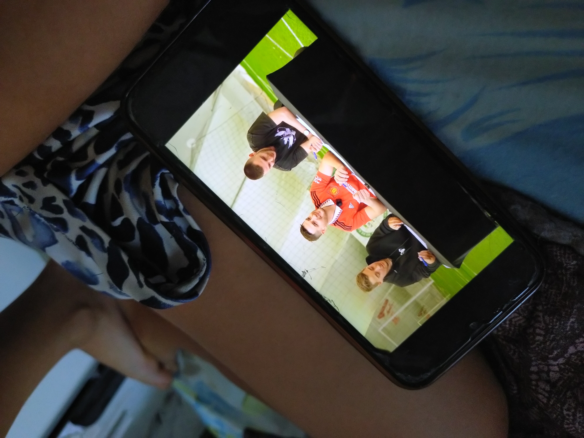 The image is a close-up, tilted shot of a smartphone screen displaying a video of three young men in what appears to be an indoor sports facility with a green turf floor marked with white lines, resembling a soccer field. On the screen, the man on the left is wearing a black hoodie and writing on a document. The man in the center is wearing a red Manchester United jersey with a white collar and also writing on a document. The man on the right is wearing a black t-shirt and a watch, and is also writing on a document. The background on the screen shows a white netted wall. The smartphone itself is black with a cracked screen and is resting on a blue fabric surface, possibly a bed or sofa. A person's legs, partially covered by a patterned fabric with blue and white markings, are visible in the foreground and to the right, obscuring parts of the smartphone and the background. The overall scene suggests a casual indoor setting, likely a home, with someone watching a video on their phone. There is no visible indication of the city of Stara Zagora, Bulgaria.