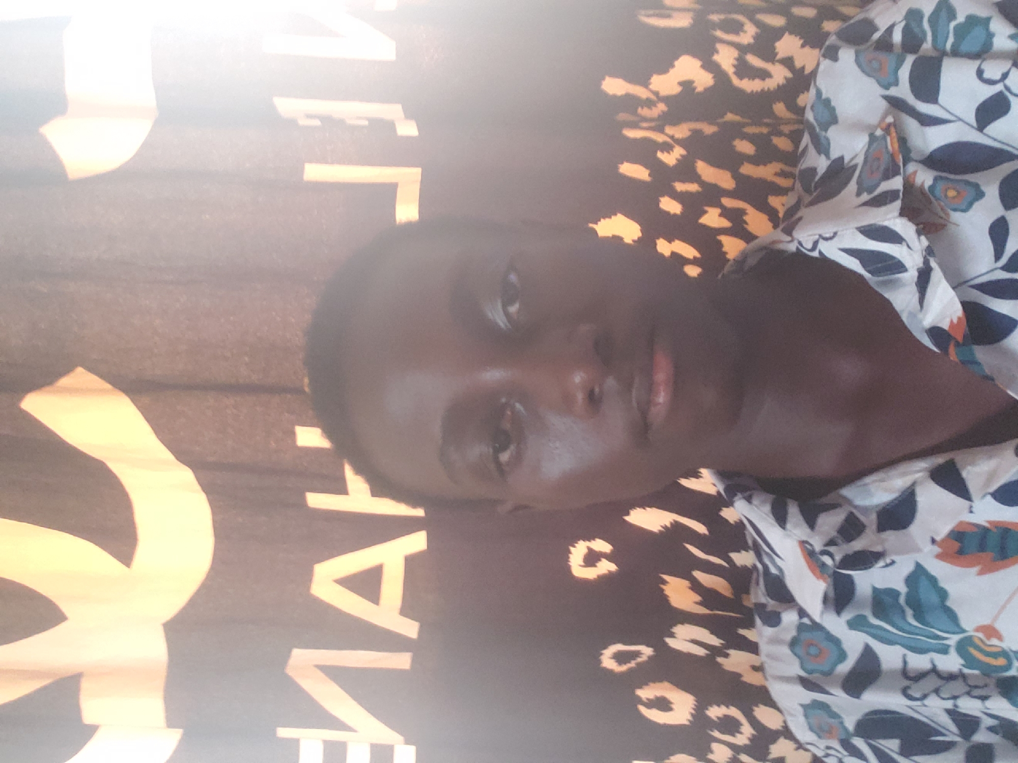 A dark-skinned person is lying on a patterned surface. Part of the surface is a black background with gold leopard print, and another part has tan writing. The person is wearing a shirt with a floral print in shades of blue, white, and orange. The location is Alalubosa, Nigeria.