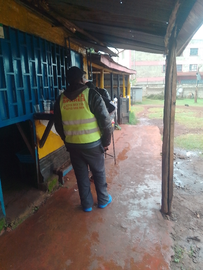 The image shows a man standing in front of a shop in Gitaru, Kenya. The man is wearing a yellow vest with a red inscription, "CYCLE SPARES," with two phone numbers, "0715 353 333" and "0712 888 888." He is also wearing blue shoes and black pants. The shop has a blue door and a sign above it. There is another person partially visible in the image. The scene is partially covered by an awning, and the ground is wet with water. The environment appears to be rural with a dirt ground and a small building in the background.