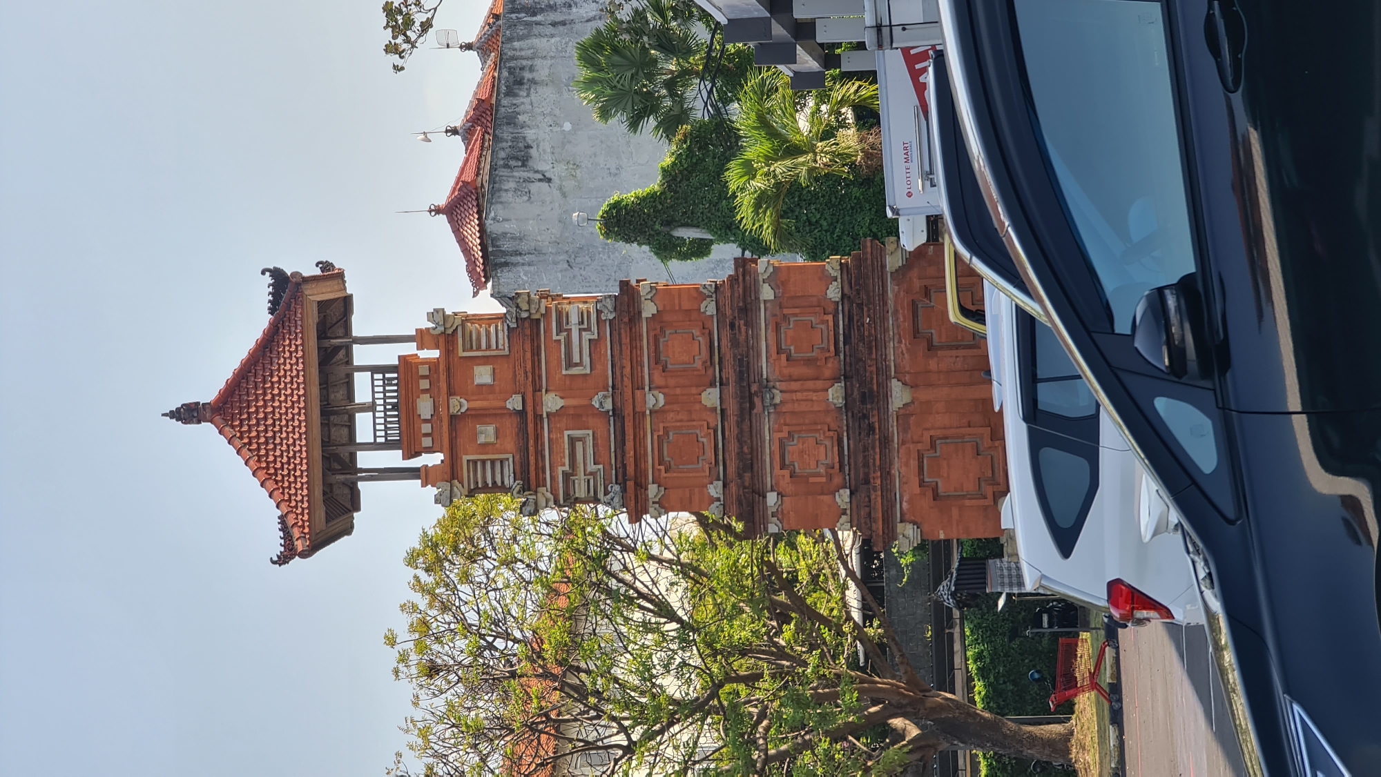 A tall, multi-tiered orange brick tower with a red tiled roof stands prominently in the center of the frame. The tower features intricate decorative patterns on each level, with stacked stone elements. To the left of the tower, a large tree with green foliage casts shadows. Behind the tower, a white building with a grey, textured facade is visible, topped with a red tiled roof. Several palm trees and dense green vegetation are also present. In the foreground, several cars are parked, including a white SUV and a black car. The scene appears to be a street or parking area in a city. A red shopping cart is visible on the left side of the frame. The sky is clear and blue. The image appears to have been taken in Denpasar, Indonesia, given the architectural style of the tower.