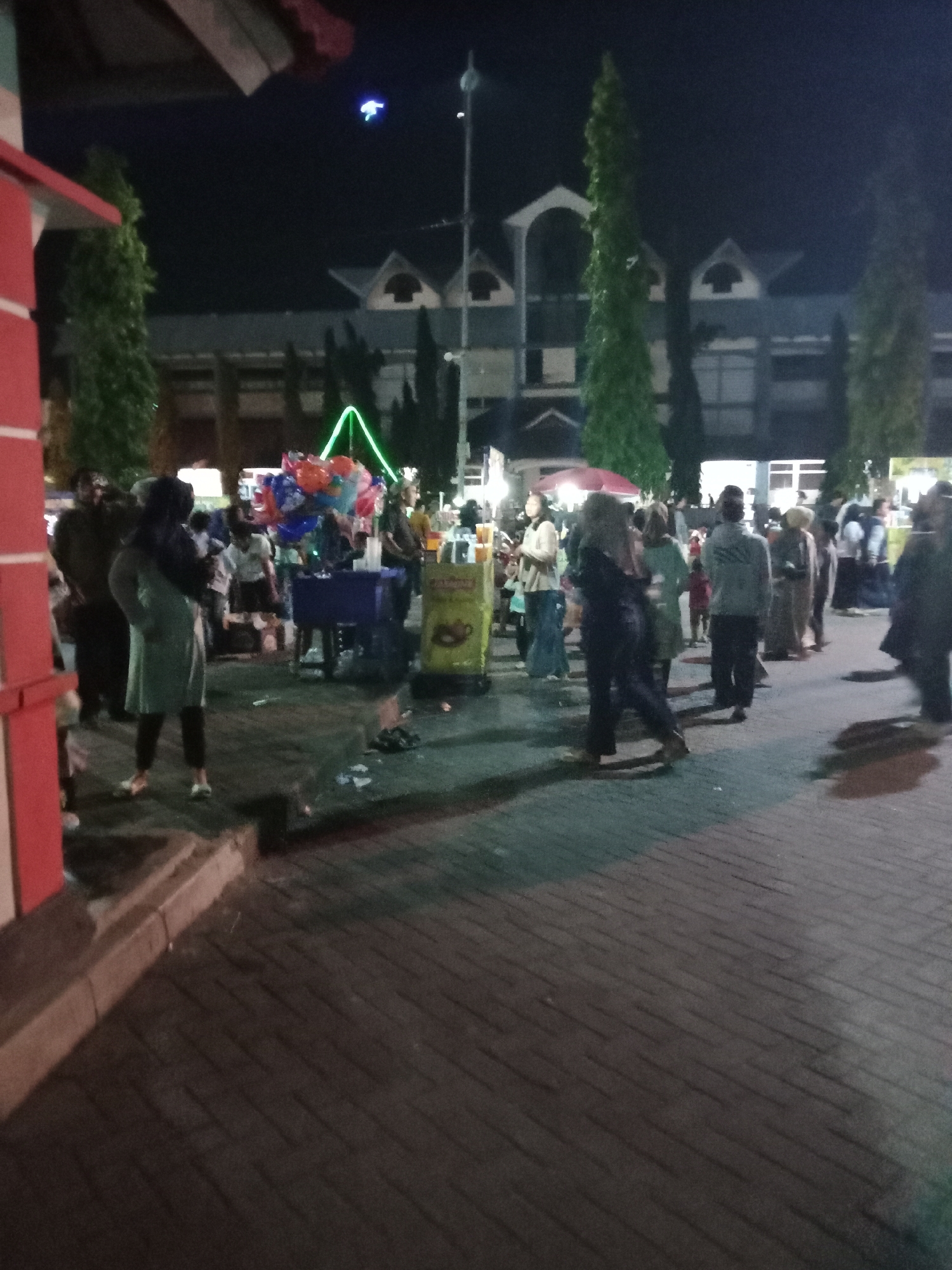 The image captures a nighttime scene in the city of Kendal, Indonesia, likely on a street or plaza, given the presence of a paved pedestrian area with interlocking bricks. The setting appears to be an outdoor market or gathering, indicated by various stalls and a crowd of people.
In the foreground, a person in a light green long-sleeved shirt and dark pants stands with their back partially to the camera, their hands clasped behind their back. To the left, a red and white structure, possibly a building or stall, is visible.
The middle ground is populated by several individuals and stalls. A blue table is set up, laden with balloons of various colors, including red, blue, and orange, some of which are tied together. A person in a light blue shirt is seen behind this table. Another stall features a yellow cart with text and graphics, likely selling food or drinks. Several other people are milling about, some interacting and others walking. One person in a dark outfit is seen walking away from the camera.
In the background, a large building with a multi-tiered roofline and arched windows is dimly lit. Tall, slender evergreen trees are interspersed in front of the building, framing the scene. The sky is dark, indicating it is night, with a faint blue light visible in the upper left portion of the image. The overall atmosphere suggests a lively event or commercial activity taking place after dark. The ground is a dark, patterned pavement, with some litter scattered around.
