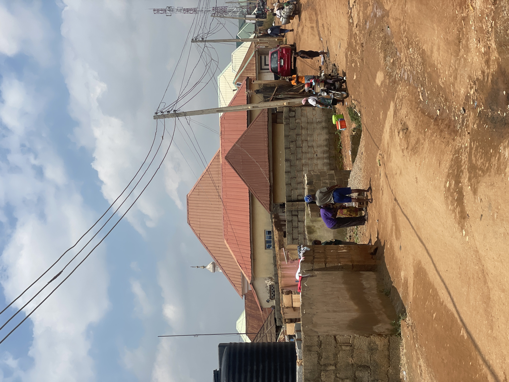 This is a photograph taken in the city of Kurunduma, Nigeria, under a cloudy sky. The scene depicts a street with a reddish-brown dirt surface, exhibiting signs of wear and potential erosion, with some puddles and debris visible. Several buildings with red, orange, and light-colored corrugated metal roofs are present. A black water tank stands prominently on the left.
In the foreground, a section of a wall constructed from concrete blocks is visible, with a plastered section showing signs of weathering. A group of people are gathered near the wall. One individual wearing a purple shirt and another in a green shirt appear to be interacting with something on the ground. Further down the street, a red car is parked.
On the street, there are several people. Some are walking, while others are near a motorcycle. One person is pushing a cart loaded with goods, indicating commercial activity. Utility poles with numerous wires are scattered throughout the scene, suggesting the presence of electricity and possibly telecommunications infrastructure. In the distance, a cellular tower is visible against the sky. The overall impression is of a busy, lived-in urban environment in Nigeria.