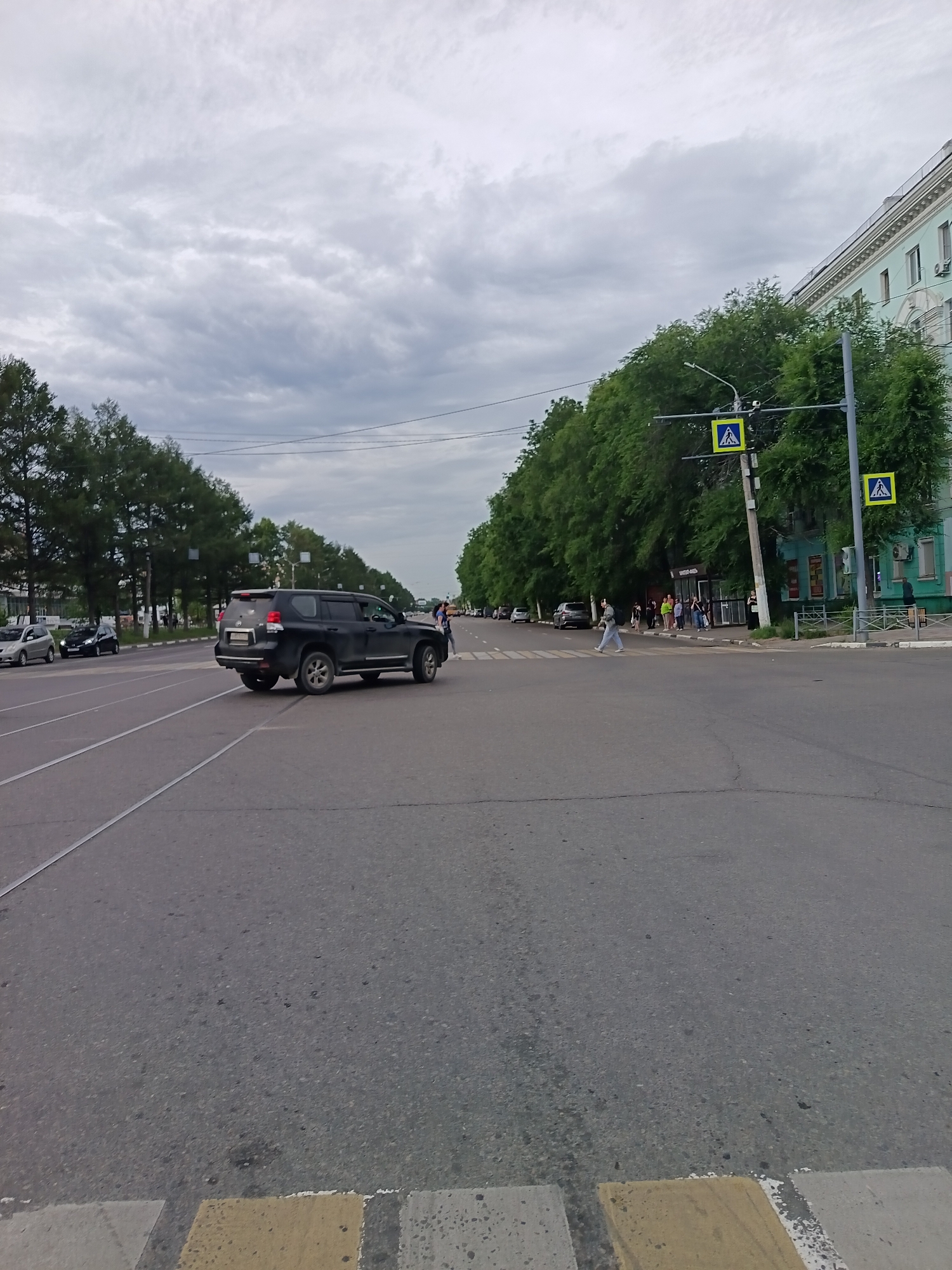 The image depicts a street scene in Komsomolsk-on-Amur, Russia, under an overcast sky. A black SUV is traveling across a crosswalk from left to right in the foreground, with its brake lights illuminated. To the right of the SUV, several pedestrians are crossing the street, some walking in the same direction as the SUV and others walking in the opposite direction. There are two pedestrian crossing signs visible, one on a pole to the right of the road and another mounted on a traffic light pole further down the street.
The road is asphalt with visible tire tracks and painted lane lines. On the left side of the road, a row of trees lines the street, with more trees and buildings visible in the background on the right. The buildings are multi-story structures, some appearing to be residential or commercial. A bus stop shelter is also visible near the right side of the frame, with a few people standing under it. Power lines are strung across the upper portion of the image. The overall atmosphere suggests a typical urban environment on a cloudy day.