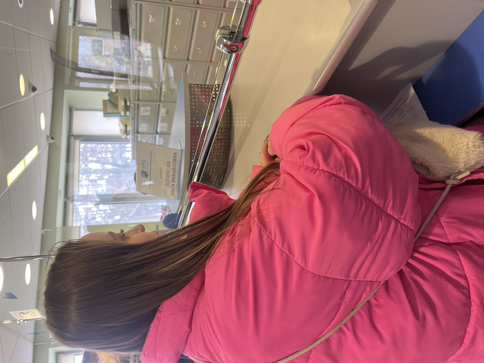 A forensic description of the scene follows:
An indoor scene depicts a young female from a side-profile perspective, facing right, at what appears to be a service counter in a public administrative office. She has long, straight, light brown hair and is wearing a voluminous, bright fuchsia (hot pink) puffer jacket. A thin, light-colored strap is visible across her back. Her right hand rests on the light-colored countertop. A polished chrome bar extends horizontally across the foreground, separating the customer area from the service area.
Behind the chrome bar and beyond a clear, reflective barrier (likely glass or plexiglass), a white sign with a blue logo (reading "КРЦ") and black text is visible. The text identifies "ЛОГИНОВА ОЛЬГА МИХАЙЛОВНА" (Loginova Olga Mikhailovna), suggesting an employee's name or service point designation. In the background, numerous rows of grey metal filing cabinets with multiple drawers are stacked, indicative of an office environment handling records. Various documents and binders are placed atop some cabinets. Large windows behind the cabinets show a view of bare tree branches, suggesting an outdoor natural environment. The white ceiling features several recessed circular lights and at least two white dome-shaped security cameras. The overall environment suggests a utility or public service center (like a Complex Settlement Center, "КРЦ") located in Chaikovsky, Russia.