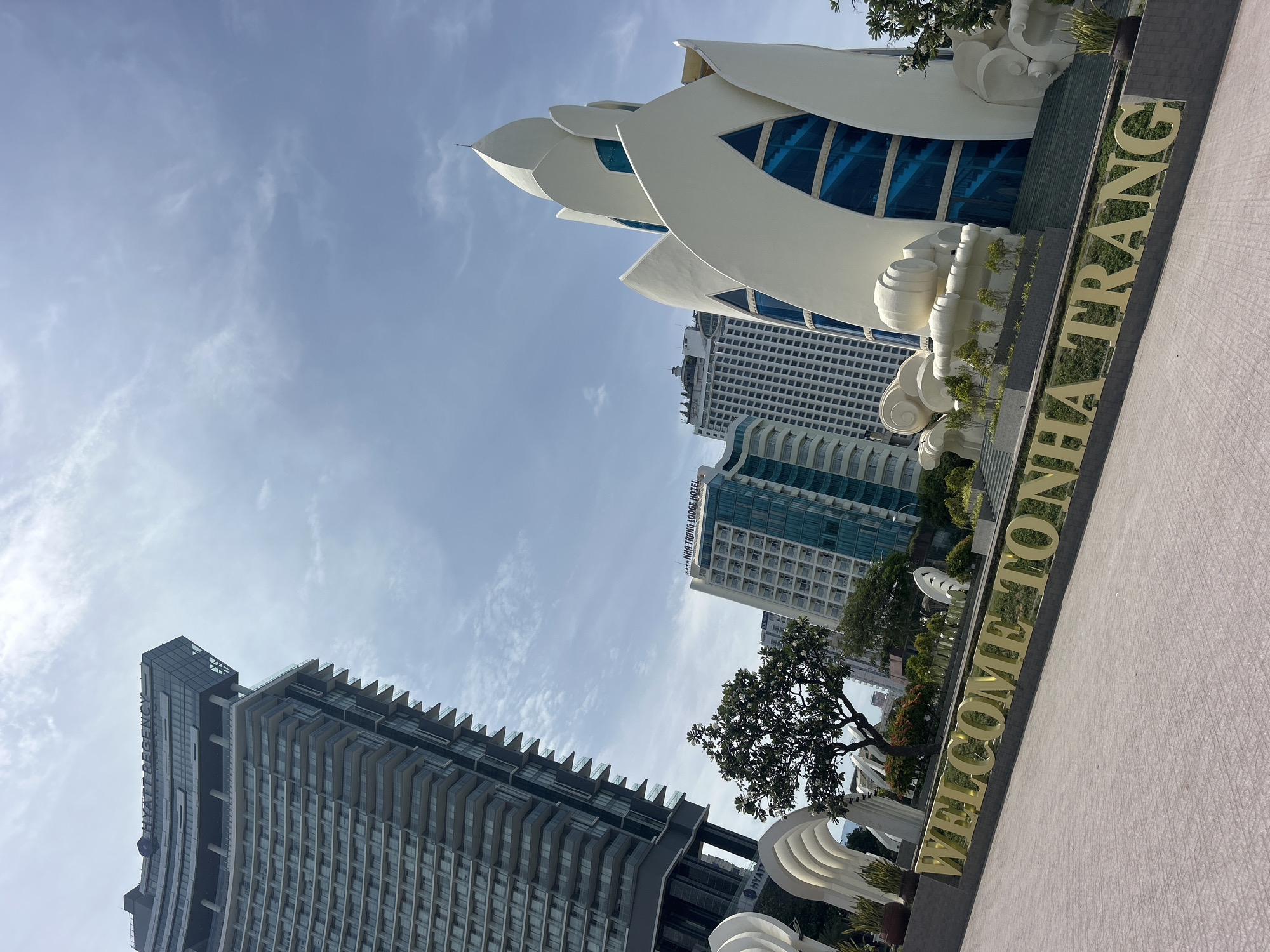 The image shows a low-angle view of buildings and a welcome sign in Nha Trang, Vietnam. In the foreground, a sign reads "WELCOME TO NHA TRANG" in gold, block letters. Behind the sign, a large, white, lotus-shaped structure stands out. In the background, several high-rise buildings can be seen, including one labeled "HYATT REGENCY HOTEL" and another labeled "NHA TRANG LODGE HOTEL." A tree is visible, positioned between the Hyatt Regency Hotel and the welcome sign. The sky is pale blue with scattered clouds. The image appears to have been taken from a public space, possibly a sidewalk or plaza.