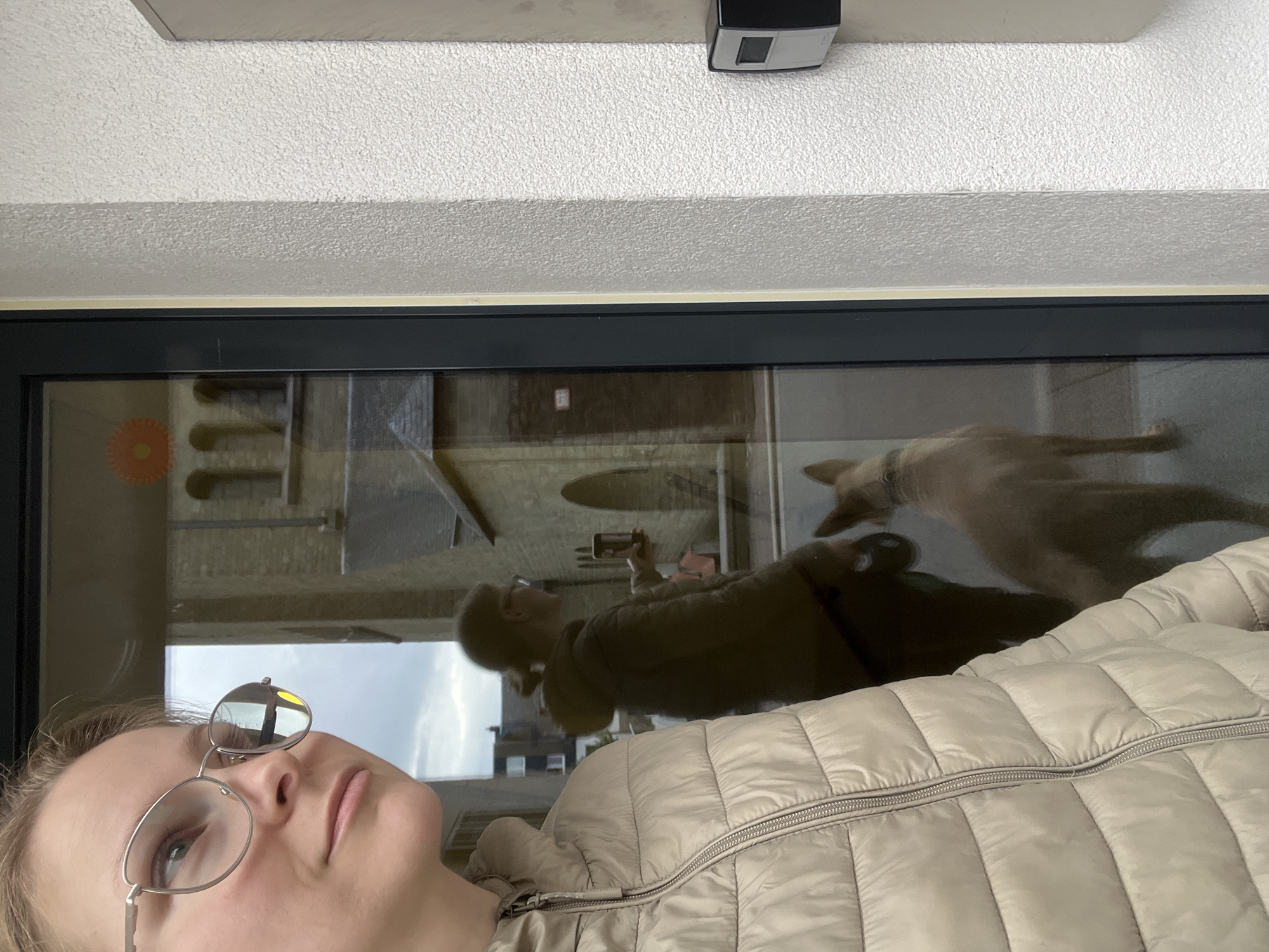 In Saint-Vith, Belgium, an individual wearing a light-colored puffer jacket and glasses is positioned in the foreground, facing left. Behind them, a reflection in a large glass window shows another individual holding a phone, a dog, and the facade of a building with arched windows, possibly a church. To the right of the glass, a textured white wall features a dark-colored intercom or access control device. The sky appears overcast.