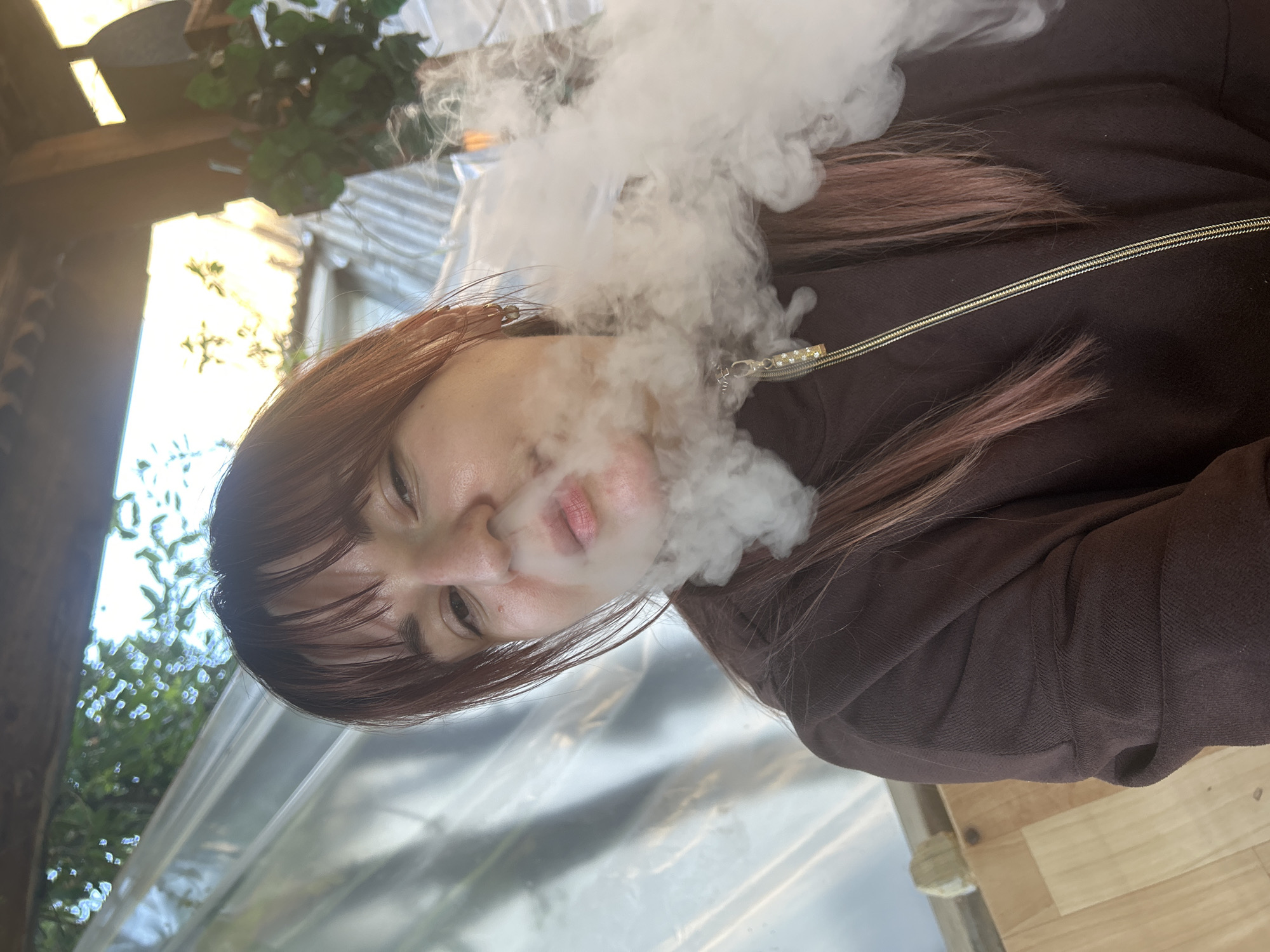A woman exhales a plume of white vapor, likely from a vaping device, directly into the camera's field of view. She is wearing a brown jacket with a visible zipper pull. Her hair is reddish-brown with pinkish highlights. The background shows a structure with a translucent corrugated roof, possibly a greenhouse or covered patio. Beyond the roof, foliage and the sky are visible. The location in Фоки, Russia cannot be verified from the image.