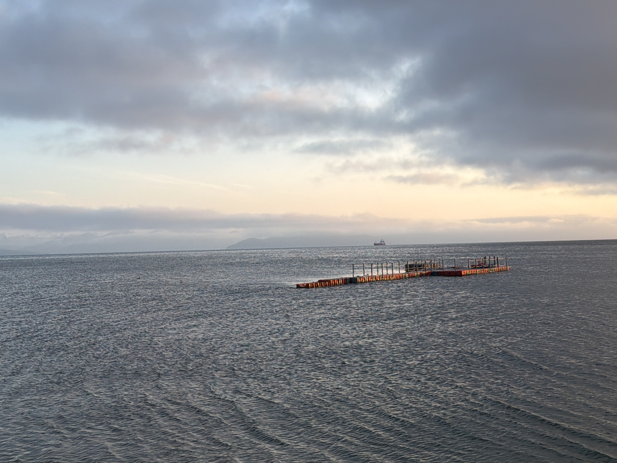 The image captures a tranquil scene of the Avacha Bay in Petropavlovsk-Kamchatsky, Russia, under a cloudy sky transitioning towards sunset. The water is calm, with gentle ripples reflecting the muted light of the sky, which exhibits soft hues of grey, blue, and peach. In the distance, a faint outline of mountains can be seen along the horizon, suggesting a coastal or island landscape.
A prominent feature in the middle ground is a floating platform or dock structure, its red base contrasting with the grey water. The platform is equipped with orange railings or barriers, and appears to be composed of modular sections. There are no people visible on the platform or in the immediate vicinity.
Further in the background, a lone cargo ship is sailing on the horizon, a small silhouette against the vast expanse of the bay and the sky. The overall atmosphere is one of quietude and vastness, typical of a late afternoon or early evening on the Kamchatka Peninsula. The image appears to be taken from a slightly elevated vantage point, possibly a pier or a nearby shore.