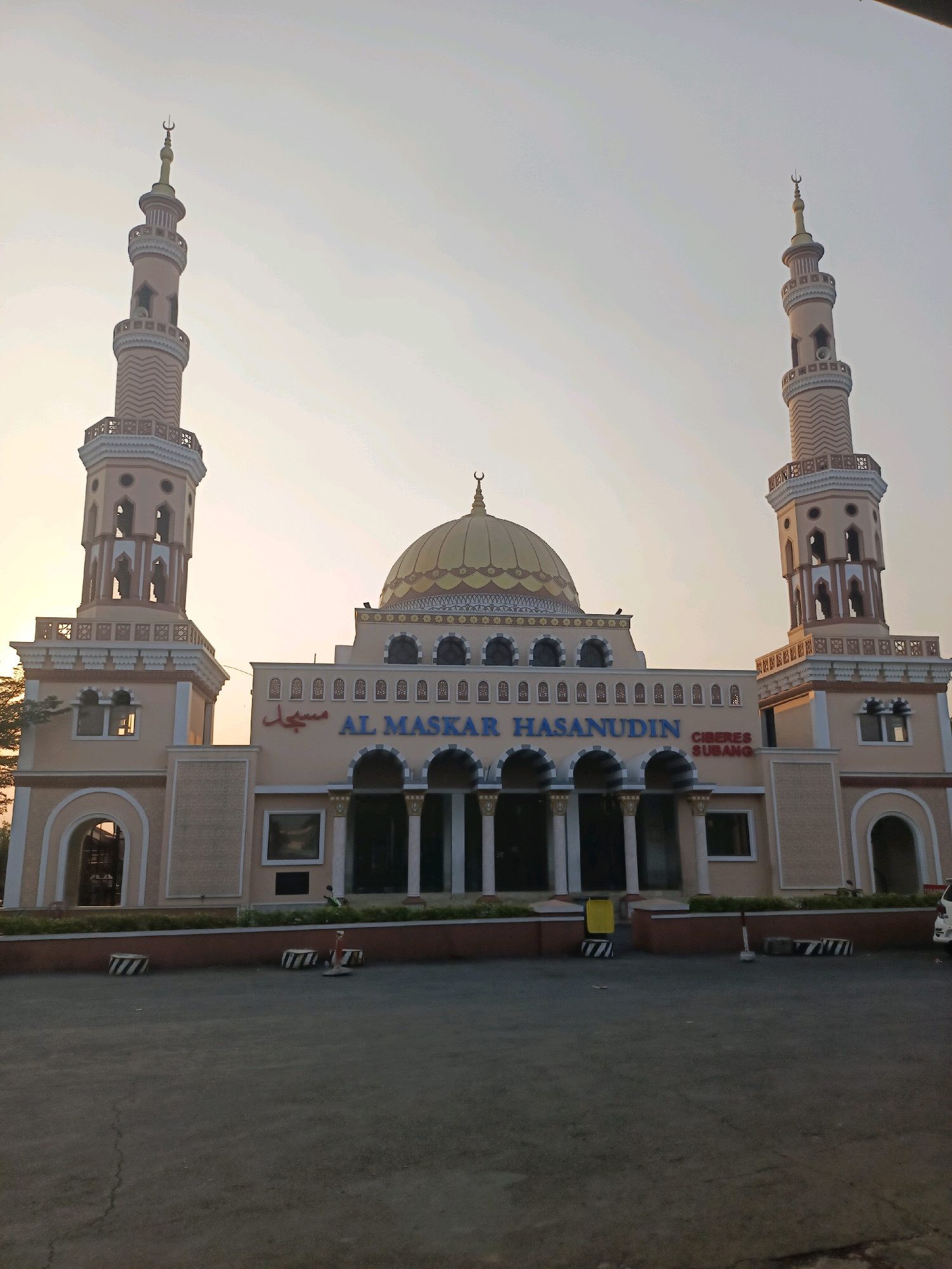 This is a front-facing, eye-level, medium shot of the Al Maskar Hasanudin Mosque in Ciberes, Subang, Indonesia. The mosque is a large, tan building with two prominent minarets, a golden dome, and a grand entrance with arches and columns. The text "AL MASKAR HASANUDIN" is written in blue letters above the entrance, and "MASJID" is written in red Arabic script to the left. The building is illuminated by the warm light of the setting sun, which casts a soft glow on the facade. The foreground is a paved area with some decorative planters and bollards. A single person is visible in the lower left corner, walking towards the mosque, and another person is standing in front of the building. A yellow trash can and some traffic cones are also present in the foreground. The sky is a pale orange and yellow, typical of a sunset.