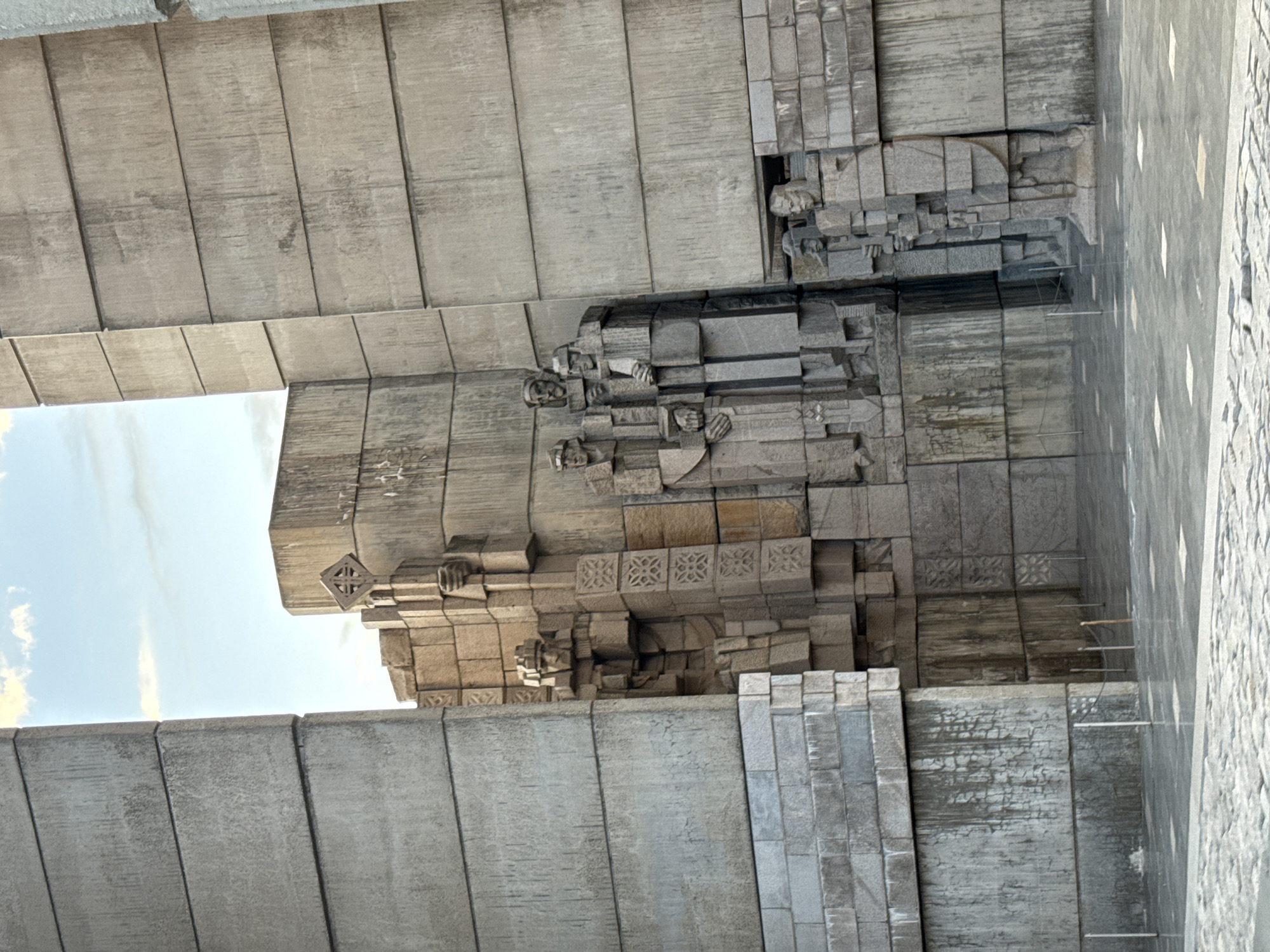The image shows a portion of the Founders of the Bulgarian State Monument located in Shumen, Bulgaria. The monument appears to be constructed primarily of concrete blocks or segments. Sculptural reliefs of figures are visible in the center of the frame. The sky is visible between the monument's concrete segments. A paved surface is located next to the monument's wall.