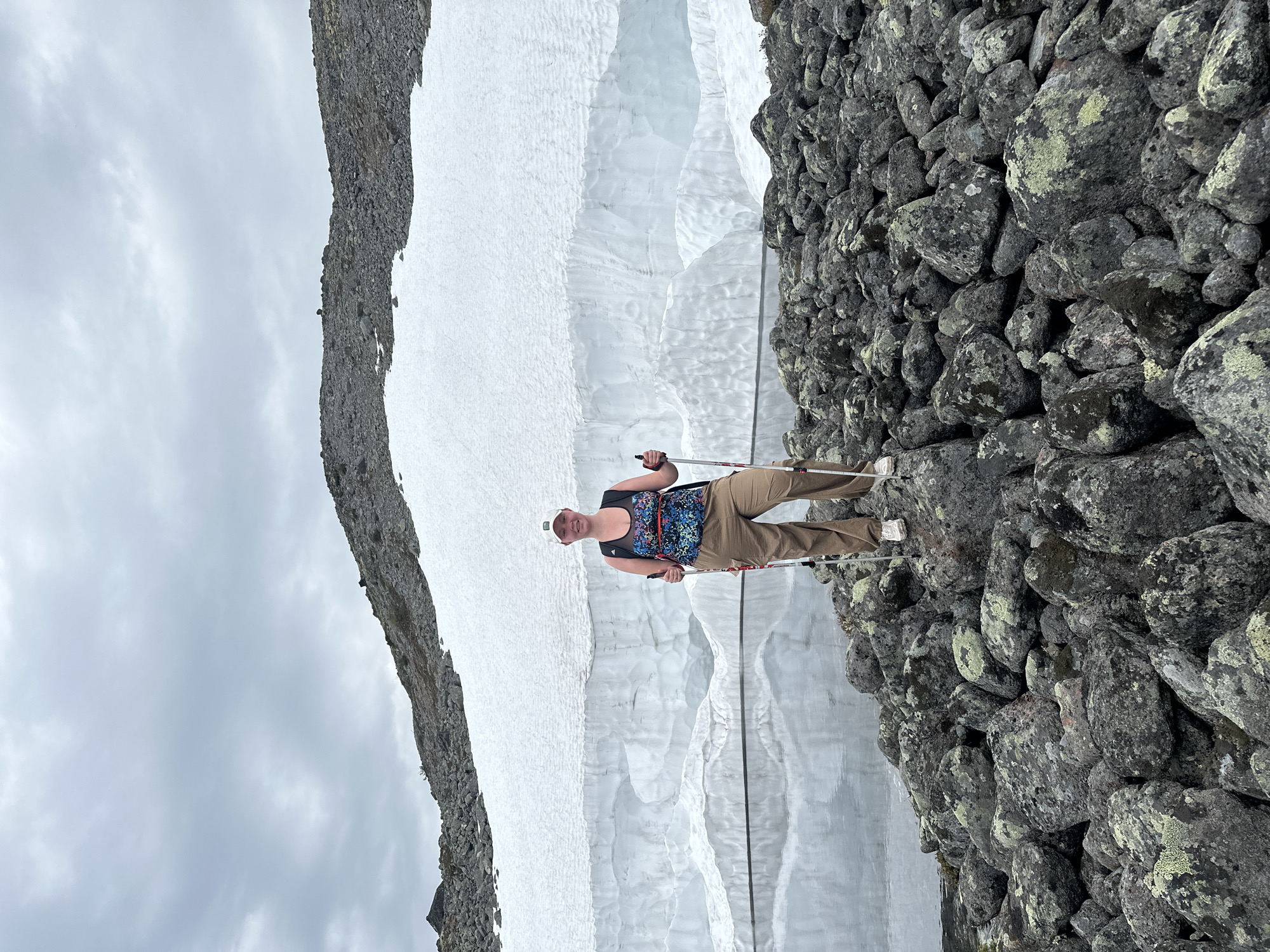 In Monchegorsk, Russia, an adult female individual stands facing the camera on an uneven field of dark grey rocks, many covered with green and yellow lichen. She is positioned in the lower-middle of the frame, holding a white and red hiking pole in each hand and smiling. She wears a white baseball cap, a patterned tank top primarily in blue, green, and black, a black mesh underlayer, light brown cargo pants, and white sneakers. A black backpack is visible on her back. Behind the individual, a calm body of water reflects a large, distinct white snow and ice wall that spans the mid-ground. The wall displays varied textures and layered formations. Above the snow and ice wall, a dark grey, rocky mountain slope with some snow patches rises towards an overcast, light grey sky.