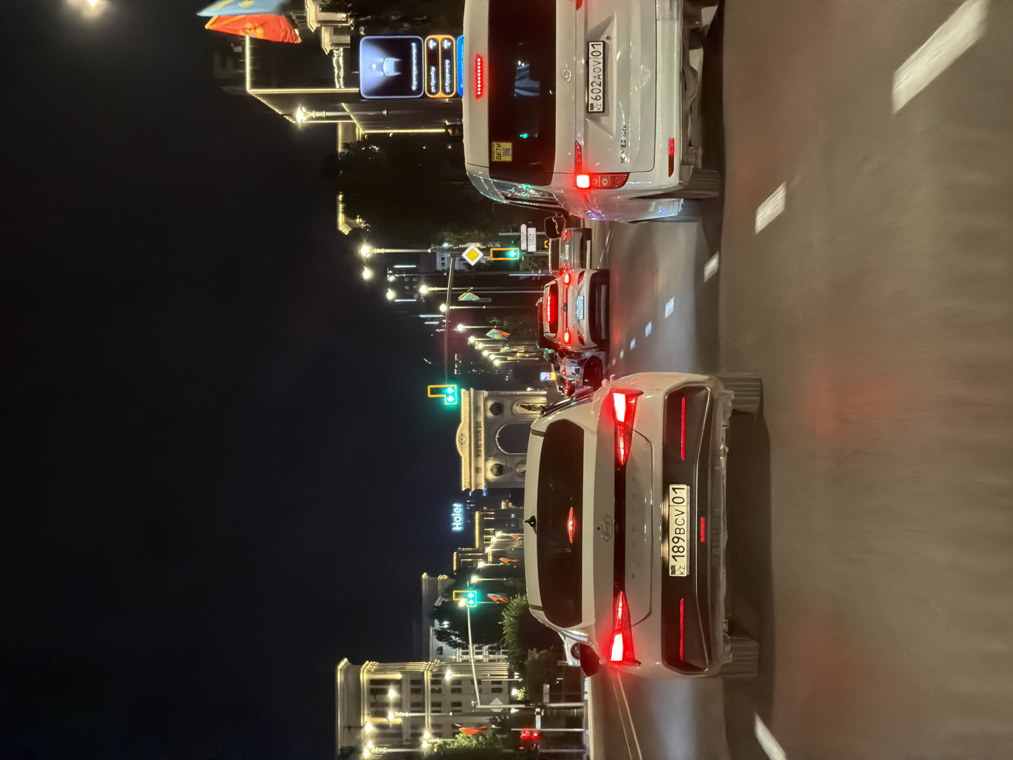 The image shows a nighttime view of a busy city street in Astana, Kazakhstan. Several cars are on the road, with their headlights and taillights illuminated. The architecture of the buildings lining the street is modern and well-lit. A prominent archway, possibly a historical landmark, is visible in the background. Traffic lights are visible, indicating the presence of an intersection. The overall scene is a typical urban environment at night, with the city's infrastructure and vehicles creating a dynamic and illuminated atmosphere.