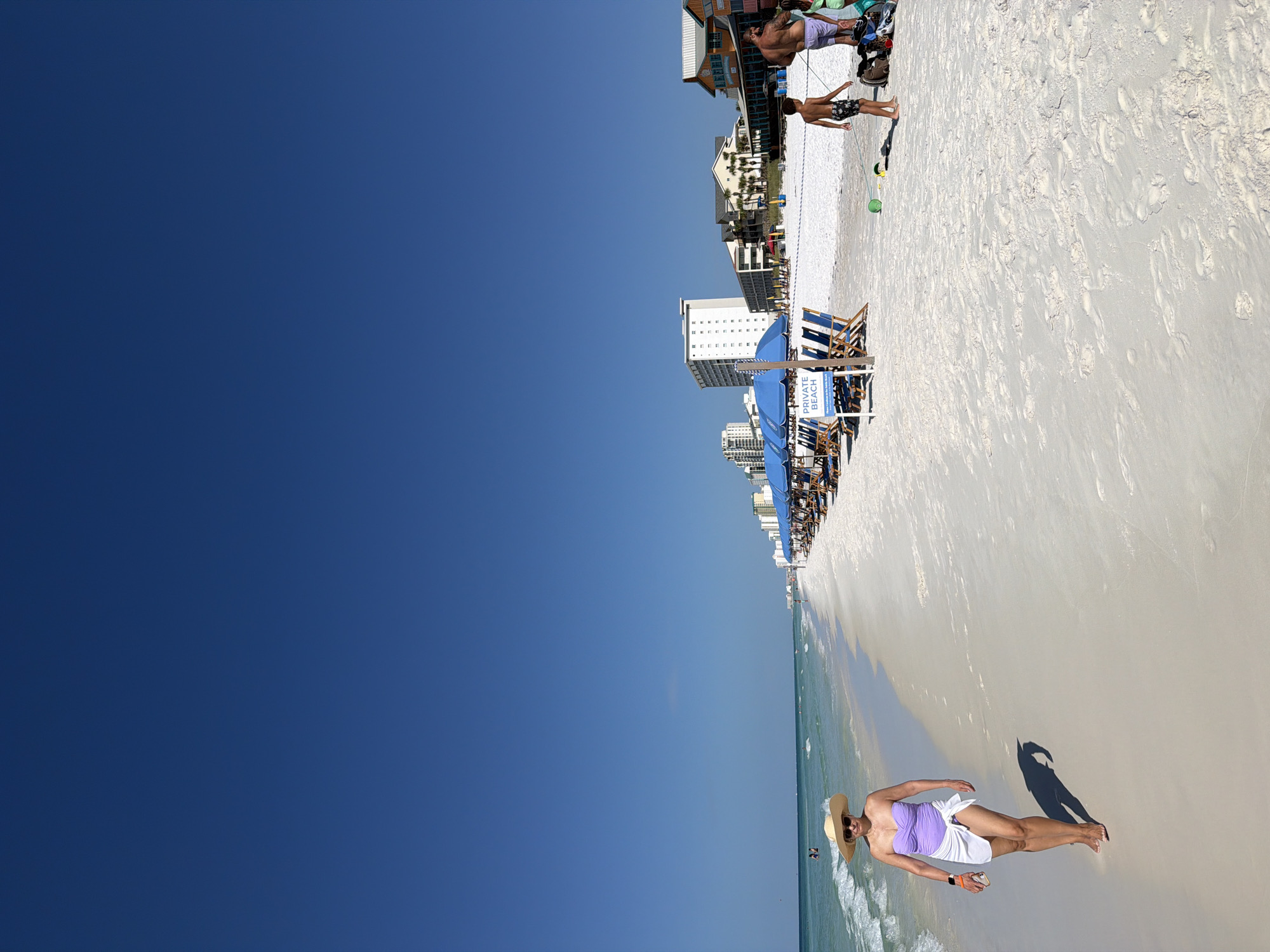 The image depicts a sunny day at a beach, likely in Destin, Florida, United States of America. The scene is divided between the ocean and the beach.
On the left, the turquoise water of the ocean is visible, with gentle waves and a clear sky above. Two figures are seen in the water, seemingly swimming.
The majority of the image focuses on the white sandy beach. In the foreground, a woman wearing a wide-brimmed straw hat, a purple strapless swimsuit, and a white sarong walks along the shoreline. She is holding a phone in her right hand and has a wristwatch on her left wrist. Her shadow is cast on the sand beside her.
Further down the beach, a row of blue umbrellas and lounge chairs are set up, suggesting a designated area for beachgoers. A sign in front of the chairs reads "PRIVATE BEACH." In the background, several multi-story buildings and what appear to be restaurants line the coast, indicating an urbanized beach setting.
To the right of the frame, a young boy in patterned swim trunks walks across the sand, pulling a green inflatable ball. Behind him, a man with a muscular build, wearing swim trunks, stands with another person, possibly a child, next to a pile of beach gear.
The overall impression is a typical beach scene with people enjoying leisure activities under a bright, clear blue sky.