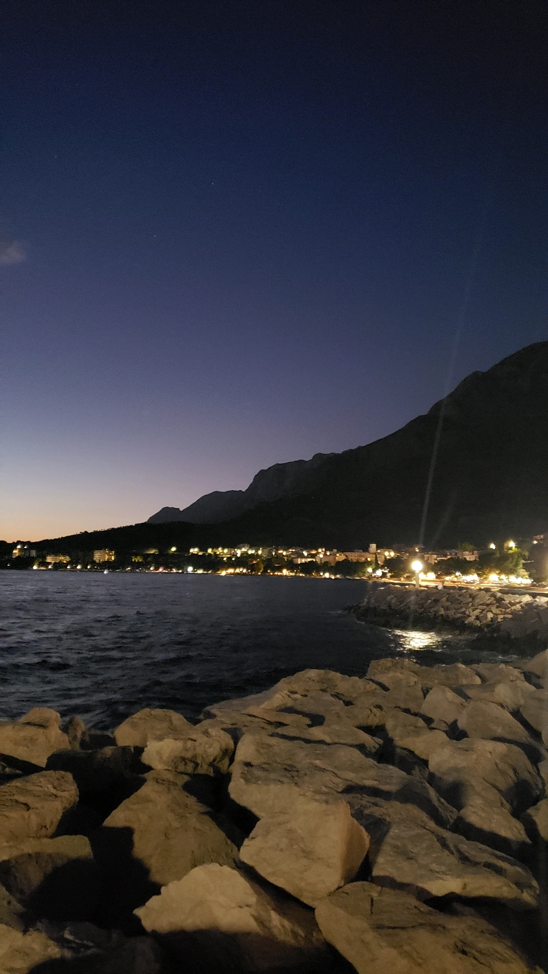 This is a nighttime image of a coastal town with a mountainous backdrop, likely taken in Općina Tučepi, Croatia, given the visual cues. The foreground is dominated by a rocky shoreline, with large, irregular stones creating a natural barrier. The dark, rippling water of the sea stretches across the mid-ground. Along the coast, a line of buildings is illuminated by numerous lights, suggesting a town or village. These lights are reflected on the water's surface. In the background, a large, dark mountain range rises steeply, its silhouette stark against the deep blue, twilight sky. A few faint stars are visible in the sky, and a faint light streak, possibly from a distant light source or lens flare, is present. The scene is tranquil, depicting a coastal settlement at dusk or after dark, with the natural landscape of mountains and sea prominent features.