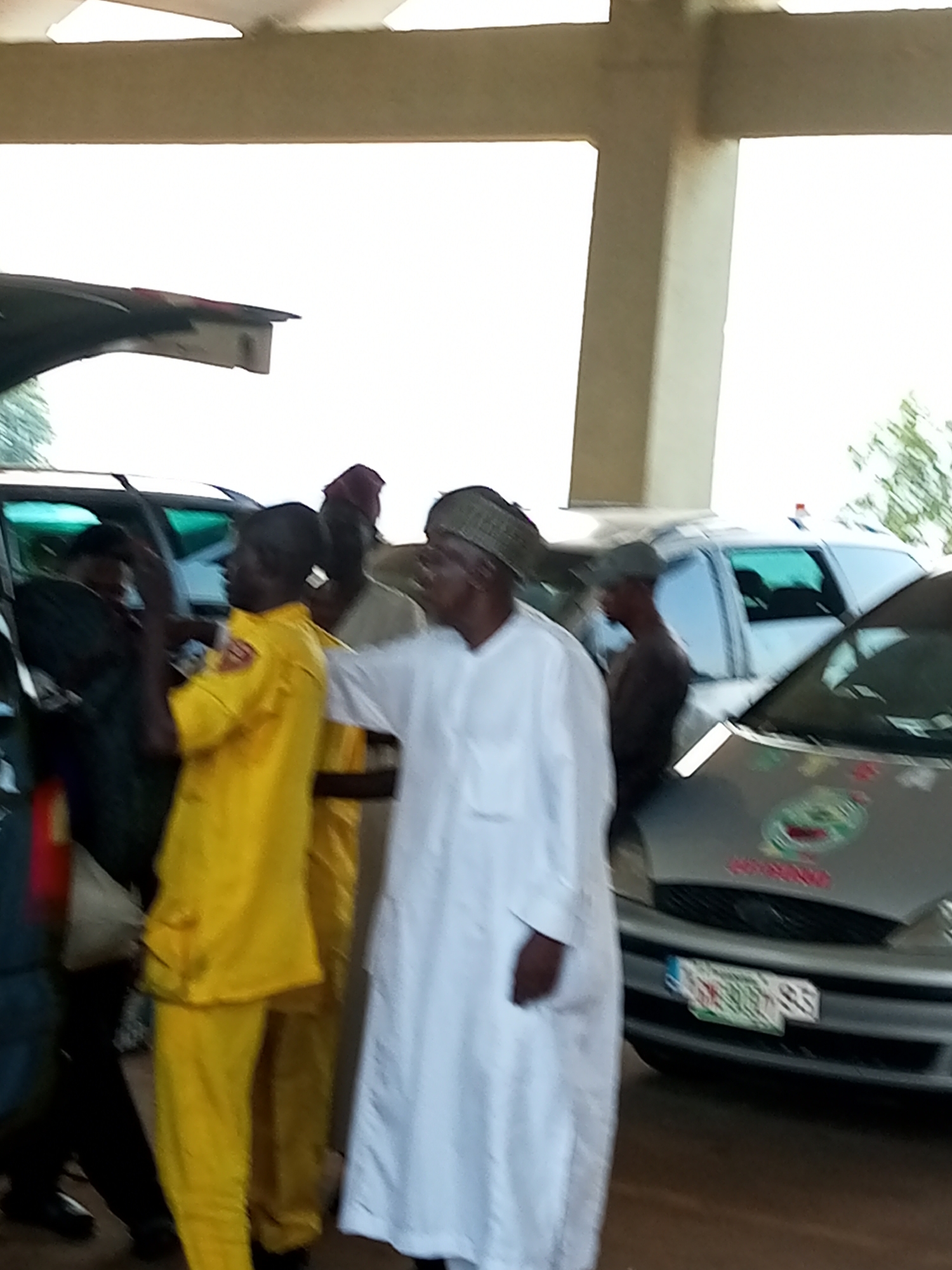 In Gombe, Nigeria, a group of men are gathered near vehicles under a covered walkway. One man, in the foreground, wears a bright yellow uniform, while another to his right is dressed in a white traditional garment and head covering. Behind them, a silver car with a logo on its hood and a license plate is visible. The setting appears to be an outdoor area with a roof overhead, possibly a parking lot or a sheltered entrance. The overall scene suggests a moment of interaction or a gathering involving these individuals and vehicles.