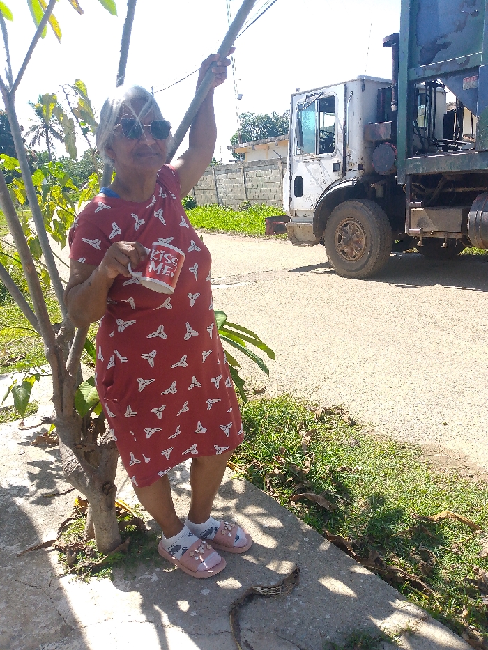 A woman in a red dress with white patterns stands next to a small tree. She wears sunglasses and holds a mug that reads "KISS ME!" Her feet are adorned with socks and pink sandals. Behind her is a large, white garbage truck on an asphalt road. A short stone wall is visible beyond the road, along with some green bushes and another building. The scene is bathed in sunlight, casting shadows on the ground.