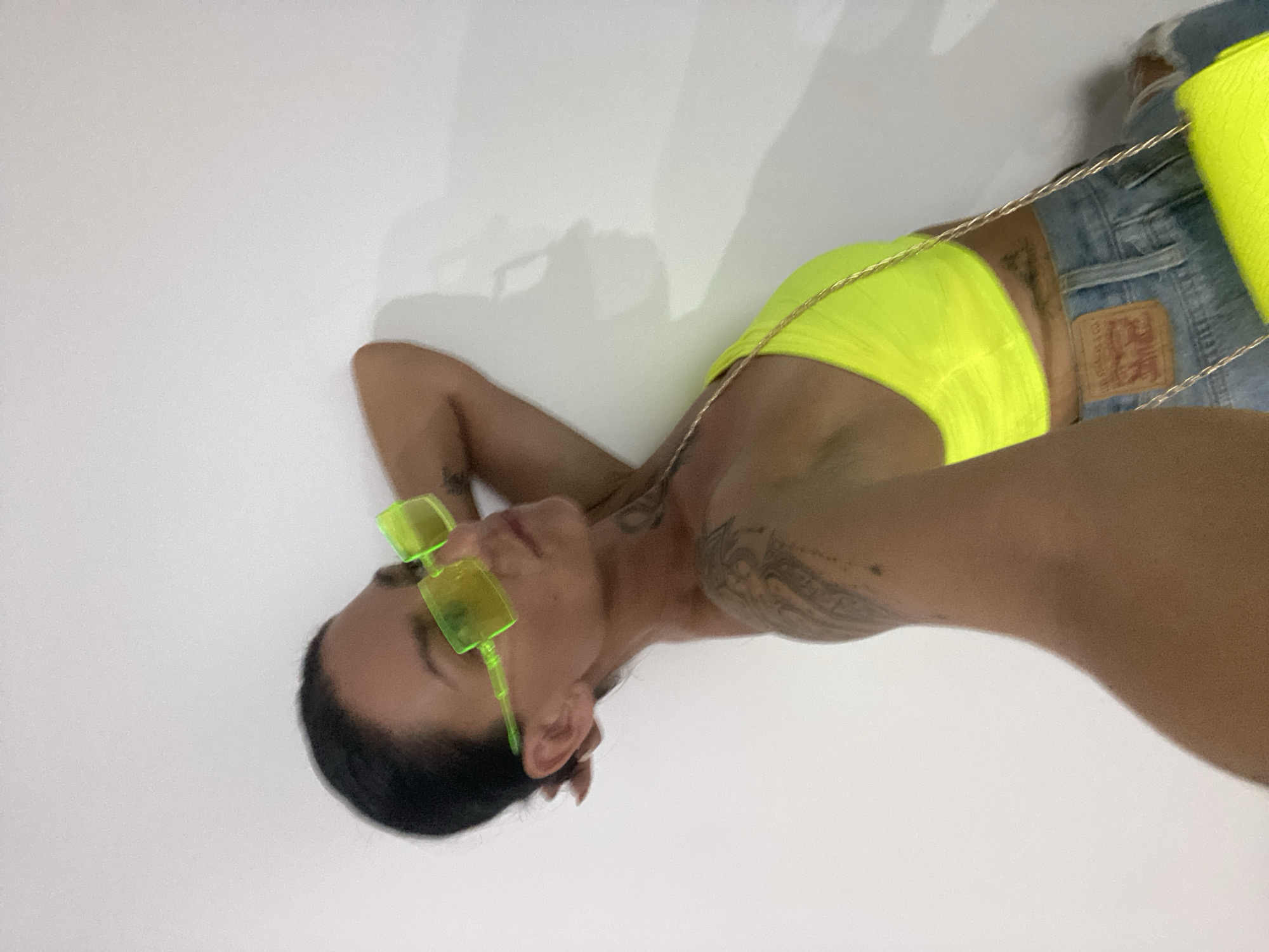 A woman is leaning against a white wall, her right arm behind her head. She is wearing neon yellow sunglasses, a neon yellow crop top, and blue jeans. A thin gold chain is visible on her left shoulder, and a neon yellow clutch is held in her left hand, partially obscuring the bottom right of the image. The wall casts a shadow to the right of the woman, creating a contrast in light and shadow. The image is a close-up of the woman's upper body, with the background being a plain white wall. The setting appears to be indoors, possibly a hotel room or a studio, in the city of Mũi Né, Vietnam.