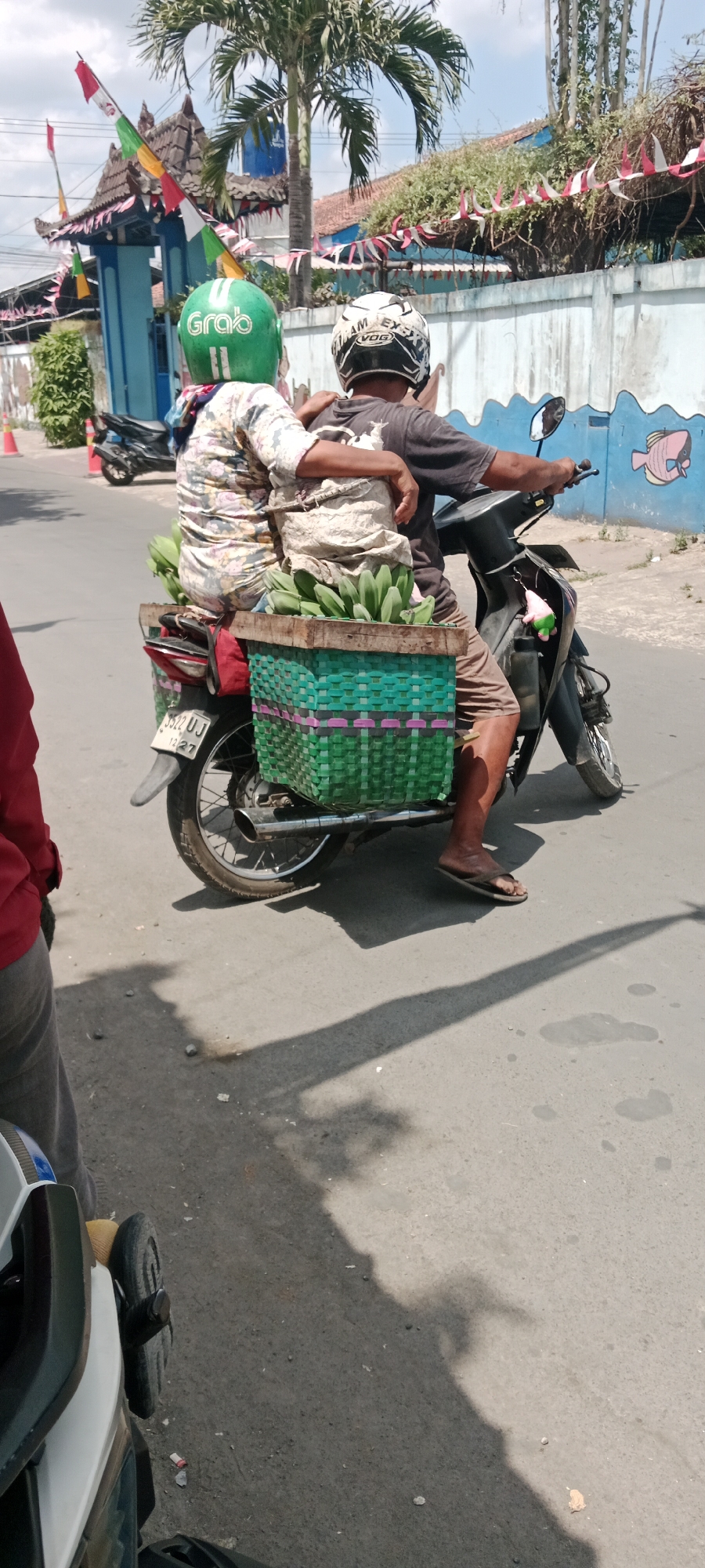 The image depicts a street scene in Purwomartani, Indonesia, likely on a sunny day given the shadows. Two individuals are riding on a motorcycle. The person in the front, wearing a green helmet with the "Grab" logo, is facing away from the camera and appears to be female, wearing a patterned shirt. The person in the back, wearing a black and white helmet, is also facing away and appears to be male, wearing a dark t-shirt. They are both embracing each other.
Attached to the back of the motorcycle is a large, green woven basket filled with bunches of green bananas. A light-colored sack is placed on top of the bananas within the basket. The license plate of the motorcycle is partially visible and reads "SJ 3522 UJ". The motorcycle itself is dark-colored and appears to be in motion, as indicated by the slight blur and the perspective of the rider.
The background features a building with a traditional Indonesian architectural style, adorned with red and white Indonesian flags and colorful banners. A palm tree is prominent behind the building. A blue wall with a white wavy design and a drawing of a fish is visible on the right side of the image. The street surface is asphalt, with some scattered debris and water marks. Another motorcycle is parked in the distance on the left side of the road. A person wearing a red shirt and gray pants is partially visible on the far left edge of the frame, also casting a shadow. The overall environment suggests a typical street in a populated area of Indonesia.