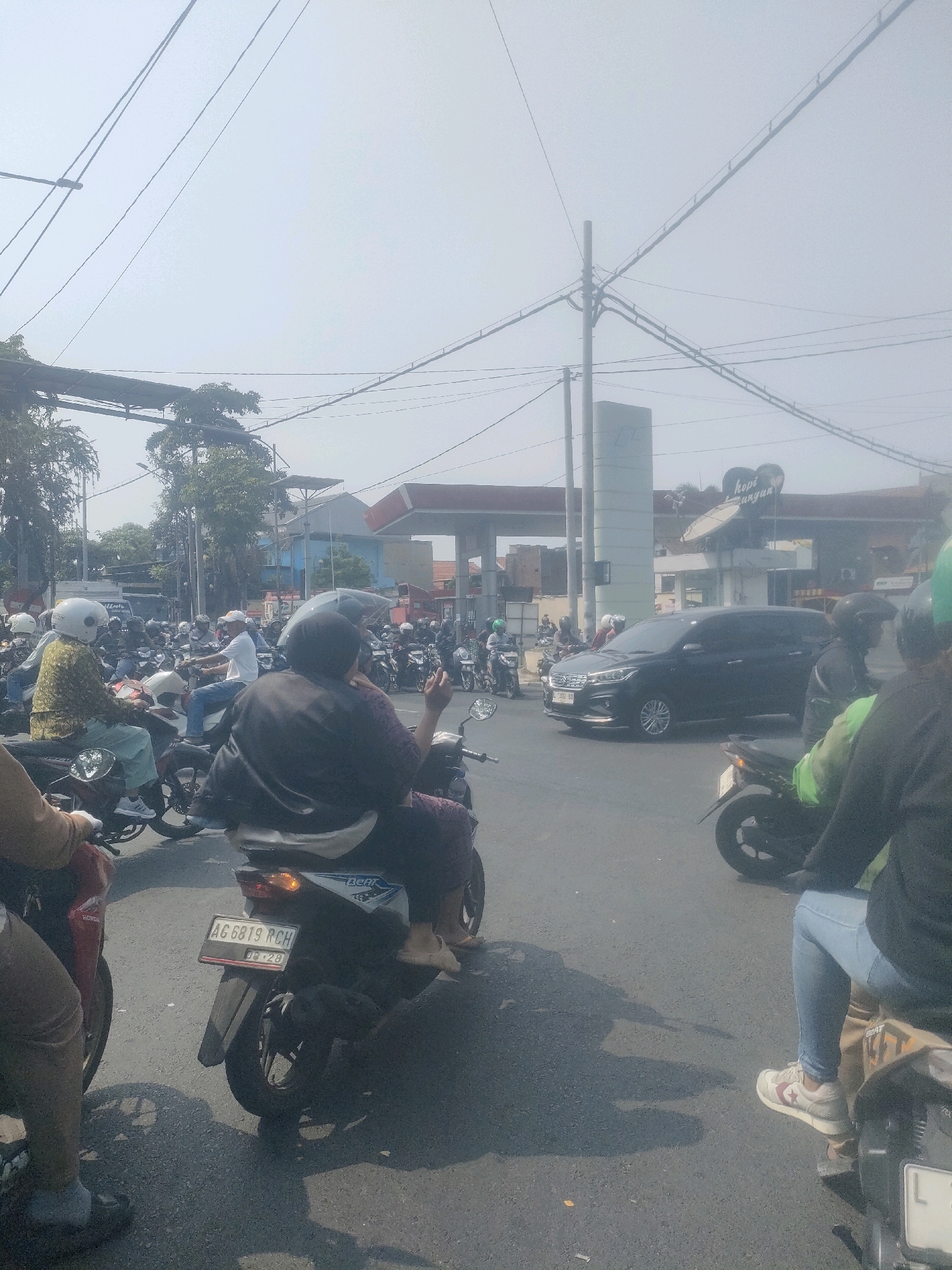 The image depicts a congested street scene in Surabaya, Indonesia, on a bright, hazy day. Numerous motorcycles, with riders wearing helmets and casual attire, are the primary mode of transportation, forming a significant traffic jam. A dark-colored MPV is visible in the middle ground, as are gas station facilities with visible signage. The environment appears to be urban, with overhead power lines and trees lining the street. The atmosphere suggests a typical busy day in a densely populated city.
