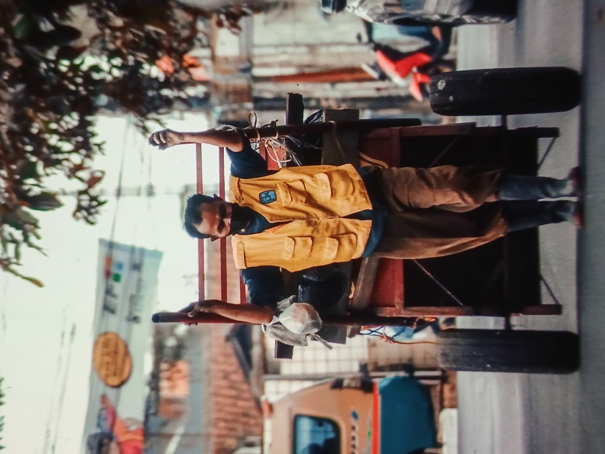 A man wearing a yellow vest and a face mask walks down a street in Setia Bakti, Indonesia, pulling a red cart. The cart has two large wheels and appears to be made of wood and metal. A bag of what looks like food is tied to the side of the cart. The man is holding onto the handles of the cart, with his arms raised. In the background, there are buildings, a van, and a motorcycle. The sky is overcast. The scene appears to be in a busy urban environment.