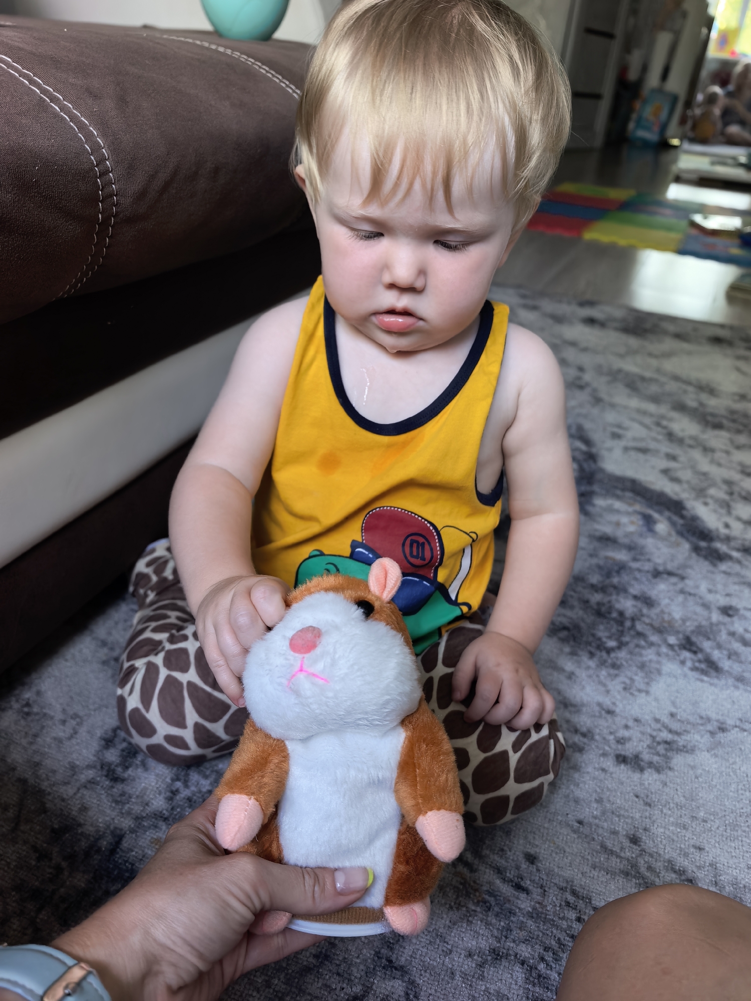 A Caucasian toddler with blonde hair sits on a rug in front of a brown couch, holding a stuffed hamster. The toddler wears a yellow tank top with a navy blue trim and a design featuring circles and cartoon figures. The hamster is brown and white with pink ears and a pink nose. The toddler's knees are on the floor, and they are wearing giraffe-patterned pants or leggings. A person's hand, with painted fingernails, is holding the hamster from the bottom. The background shows a living room area with a colorful foam play mat on the floor and a blurred view of other furniture and a window. The overall environment appears to be indoors, likely a home setting. There is no visible indication of the city of Sterlitamak, Russia.
