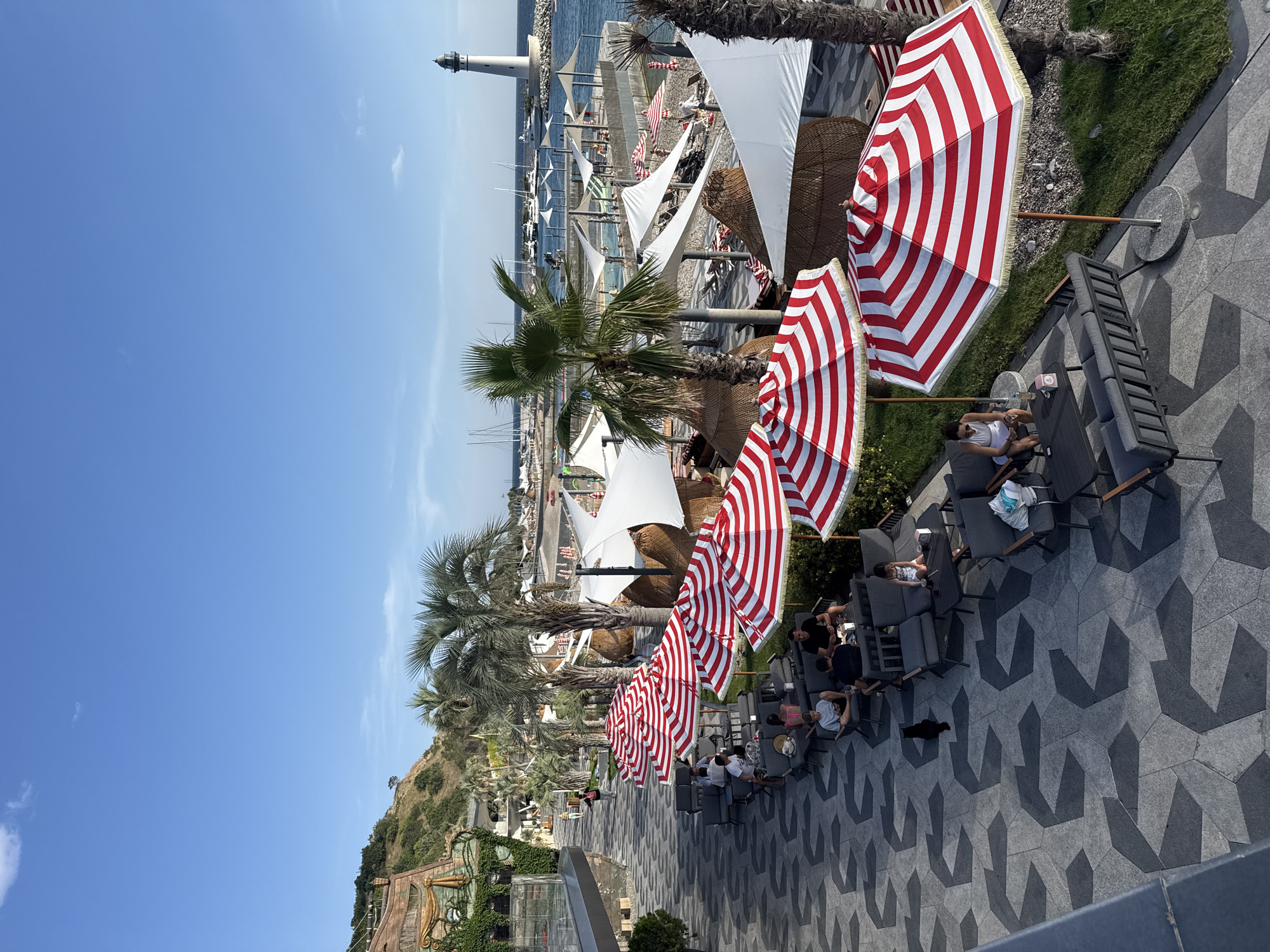 The image depicts a sunny outdoor scene at a seaside resort, likely in Simeiz, Ukraine, given the presence of the lighthouse, which is a landmark of the area. The foreground features a patterned paved walkway with several groups of people seated on dark grey outdoor furniture, some under red and white striped umbrellas. A black cat is visible on the walkway, walking away from the camera. Beyond the seating area, there is a beach with more umbrellas, thatched cabanas, and beach chairs. The middle ground shows palm trees and other vegetation, with a body of water and a pier lined with more seating and umbrellas. In the background, a white lighthouse stands on a rocky outcrop. Several sailboats and yachts are docked in the water. The sky is a clear blue with a few wispy clouds. The architecture of a building with an arched wooden facade is visible on the left side of the image, partially covered in greenery.