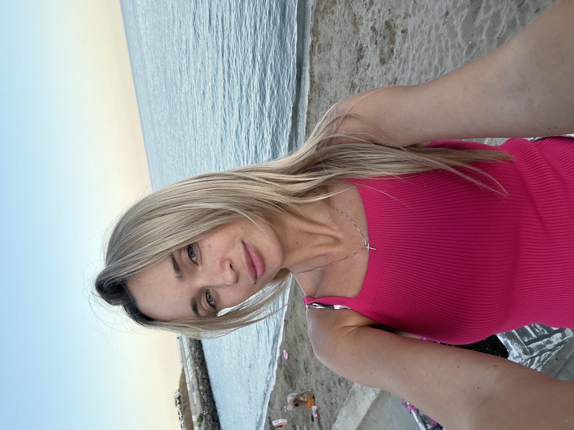 A woman with blonde hair and green eyes takes a selfie on a beach in Fontanka, Ukraine. She is wearing a bright pink tank top and a silver necklace with a cross pendant. The background shows the calm sea, a sandy beach, and a breakwater in the distance. There are a few other people visible on the beach, including one person kneeling and playing with something on the sand. The sky is clear with a soft gradient of blue and pale yellow, suggesting either early morning or late evening.