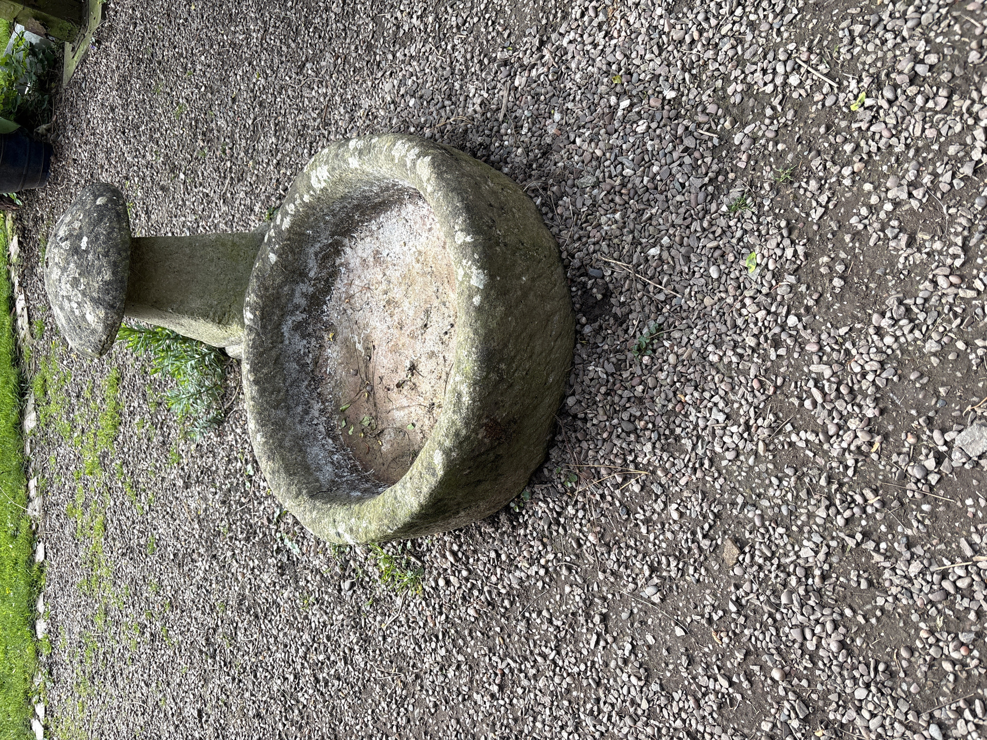The image shows a stone bird bath in Pulverbatch, United Kingdom. The bird bath, made of weathered stone, features a rounded bowl atop a shorter, narrower stone pillar. The interior of the bowl is partially filled with light-colored material, possibly sand or sediment. The bird bath sits on a bed of small, gray gravel. To the left of the bird bath, green vegetation is visible along a border. The overall setting appears to be an outdoor area, possibly a garden or yard.
