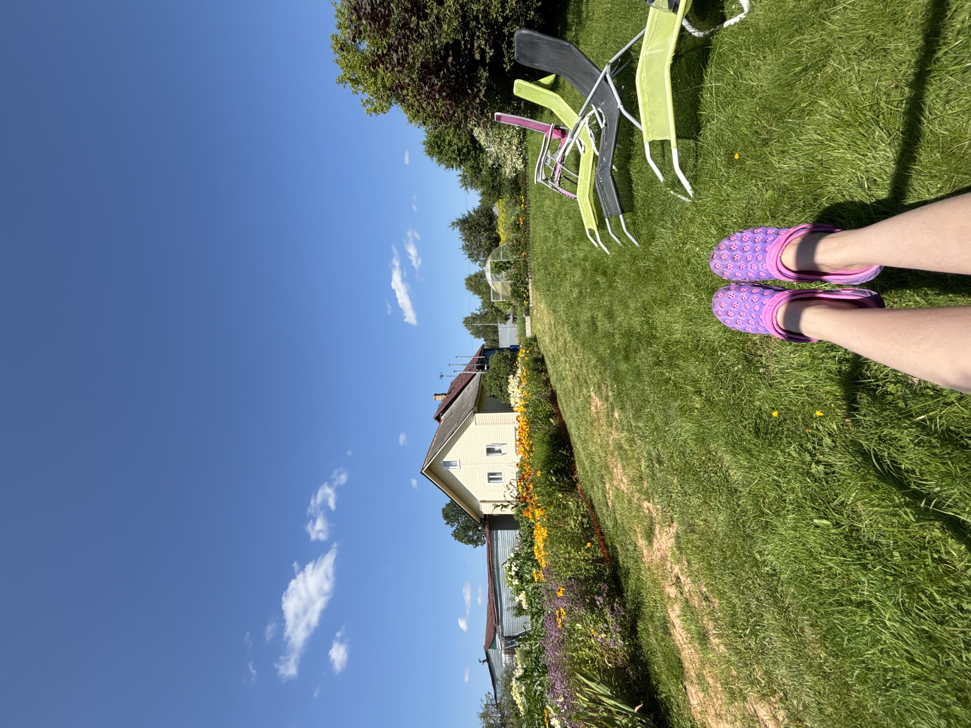 A person's legs, adorned with purple clogs patterned with small pink fruits, are visible in the foreground of this outdoor scene, positioned on a vibrant green lawn. The perspective is low, looking up towards a bright blue sky dotted with a few white, wispy clouds.
In the mid-ground, a row of colorful flowers, predominantly yellow and purple, stretches across the image, bordering a dirt path. Behind the flowerbed, a light yellow house with a red roof and a stone chimney stands under the expansive sky. Adjacent to the house is a weathered, dark-colored building with a corrugated metal roof. Further in the background, a greenhouse structure can be seen.
To the right of the frame, several lawn chairs are arranged on the grass. One is a bright pink, folded chair, with another green chair partially visible beneath it. In front of these, a black chair and another green chair are reclined, their white frames contrasting with the colorful seating. A large, dark-leafed tree dominates the upper right portion of the scene.
The overall impression is a tranquil, sunny day in a rural setting, likely a residential area in the village of Dunilovo, Russia, characterized by its traditional houses, abundant gardens, and open green spaces.
