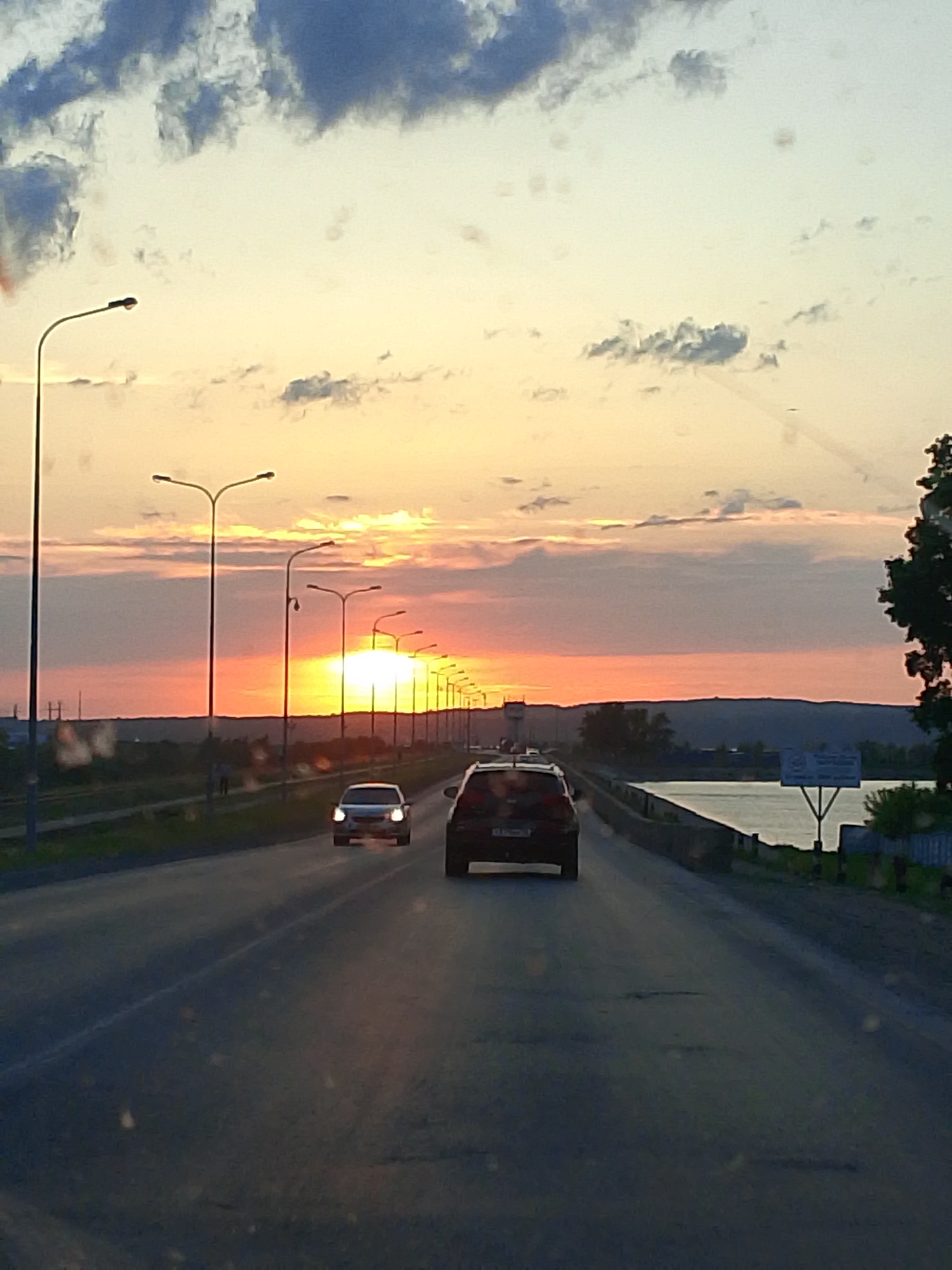 The image shows a sunset over the city of Chaikovsky, Russia. The viewpoint is from a car driving on a road with streetlights. Another car is visible in front. To the right is a body of water, potentially a reservoir or wide river. A billboard or sign is visible near the water, and hills are present in the distance. The sky is filled with clouds, illuminated by the setting sun.