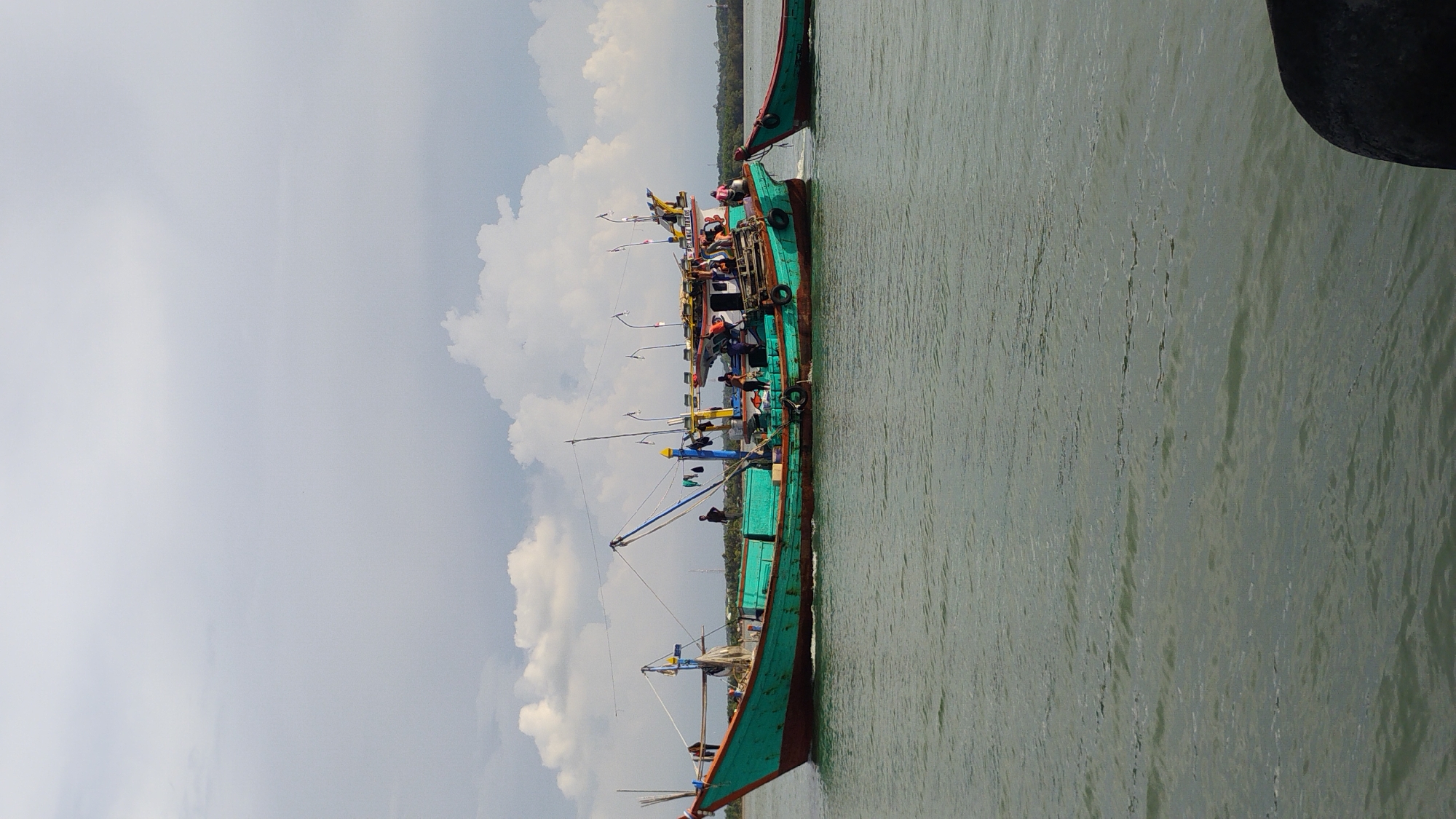 The image depicts a cloudy day over a body of water in Lhokseumawe, Indonesia. A large, green and teal fishing boat is the primary subject, positioned in the center of the frame. The boat has a red waterline and appears to be moving, as indicated by the wake behind it. Several people are visible on the deck of the boat, engaged in various activities, suggesting a working vessel. Another smaller, red boat is partially visible on the right edge of the frame, connected to the larger boat by a rope. The water is calm with subtle ripples. In the background, a treeline and structures can be discerned on the distant shore. The sky is filled with a mix of light and dark grey clouds.