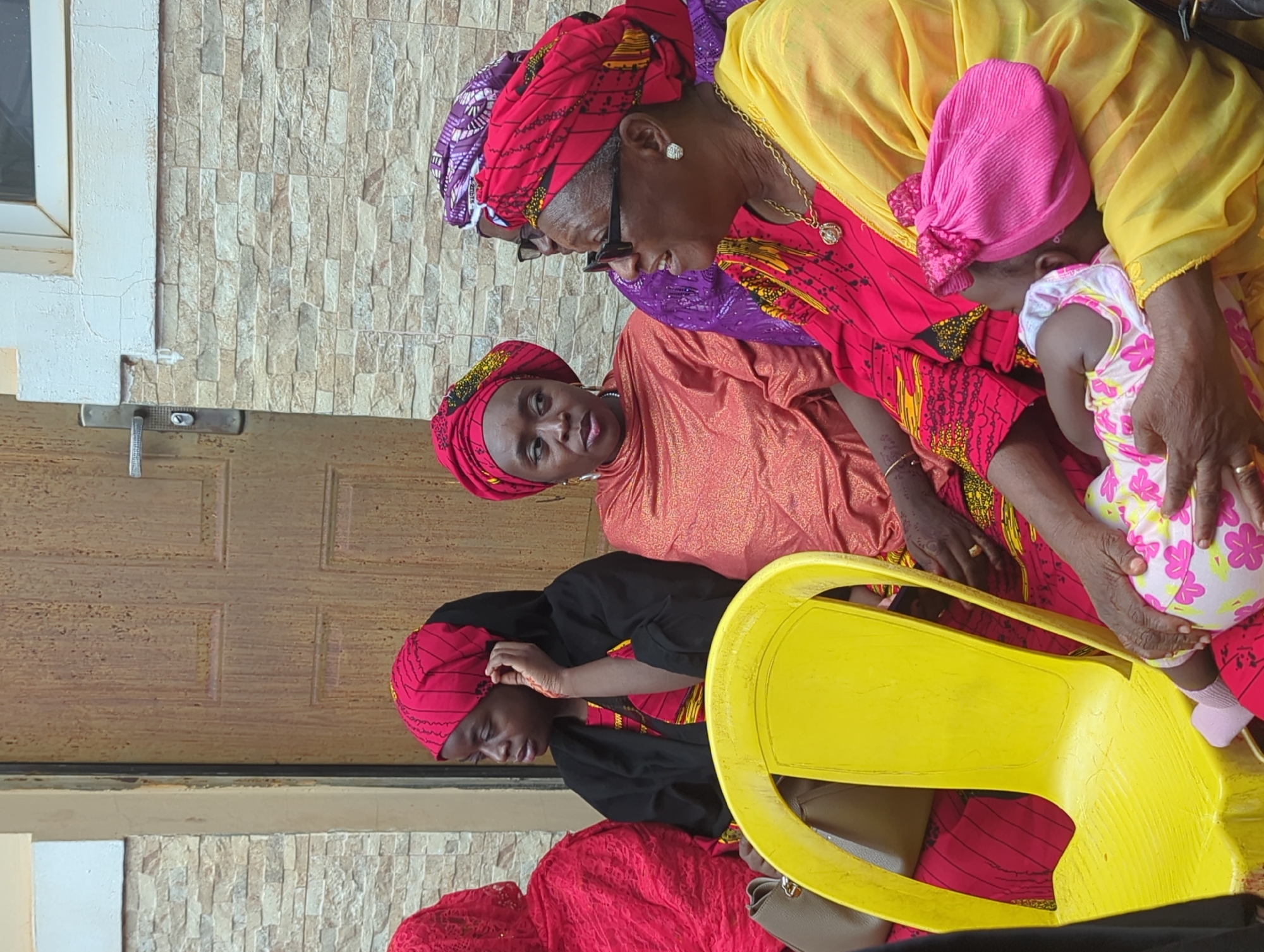 In Unguwar Rimi, Nigeria, a group of five individuals, primarily women, and one infant are seated outdoors against a building facade. On the left, a young woman, wearing a red patterned headwrap, a black outer garment over a red and yellow patterned top, sits on a bright yellow plastic chair. A grey handbag is visible beside her, and her right hand touches her ear. Adjacent, a woman in an orange headwrap and a metallic orange garment looks forward. To her right, a woman in a red and yellow patterned headwrap, wearing sunglasses and a gold necklace with a pendant, is positioned, seemingly interacting with an elderly woman. This elderly woman, adorned in a yellow headwrap, a yellow garment, glasses, and a gold necklace, holds an infant. The infant is dressed in a pink and white floral patterned romper and a pink bow-adorned hat, with its back partially to the viewer. A fifth individual, in a purple garment and headwrap, is partially obscured behind the woman in red and yellow. The background consists of a building with a lower section of light-colored, horizontally stacked stone tiles, above which a white window frame is visible on the right. A brown wooden door with a silver handle is prominent in the center-left background. The scene is illuminated by natural daylight.