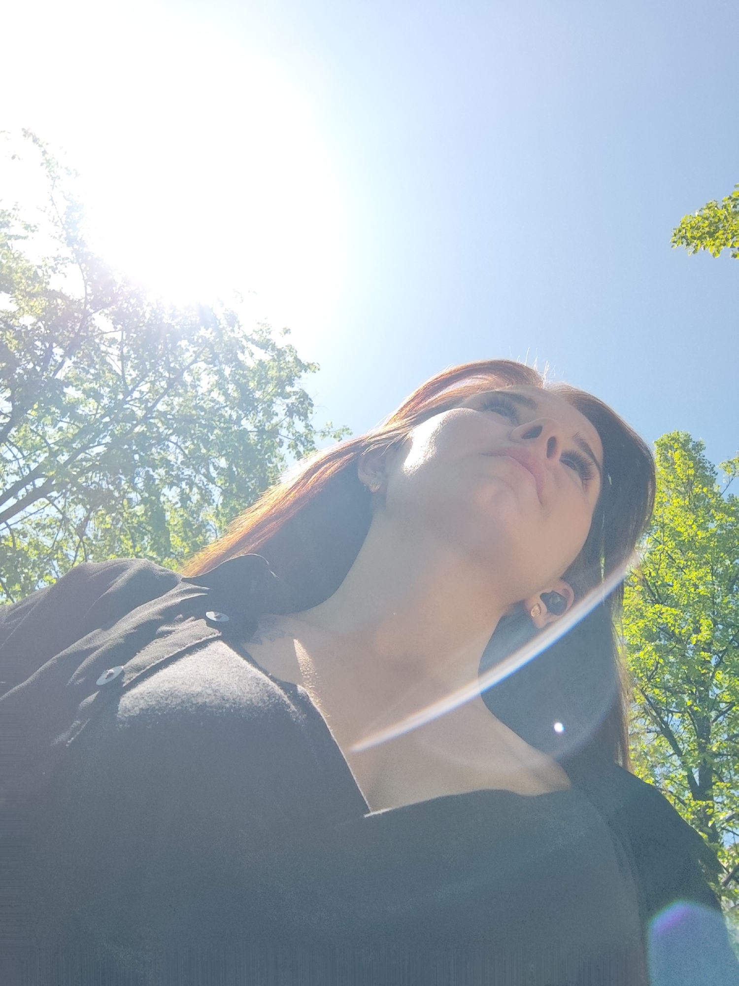 The image shows a low-angle view of a person looking upwards into a bright, sunny sky. The person has long, brown hair, and is wearing a dark-colored shirt with buttons. The sun is visible in the upper portion of the image, causing lens flare. Green foliage of trees is visible at the periphery of the image. There is insufficient information to identify any specific location in the city of Chaikovsky, Russia.