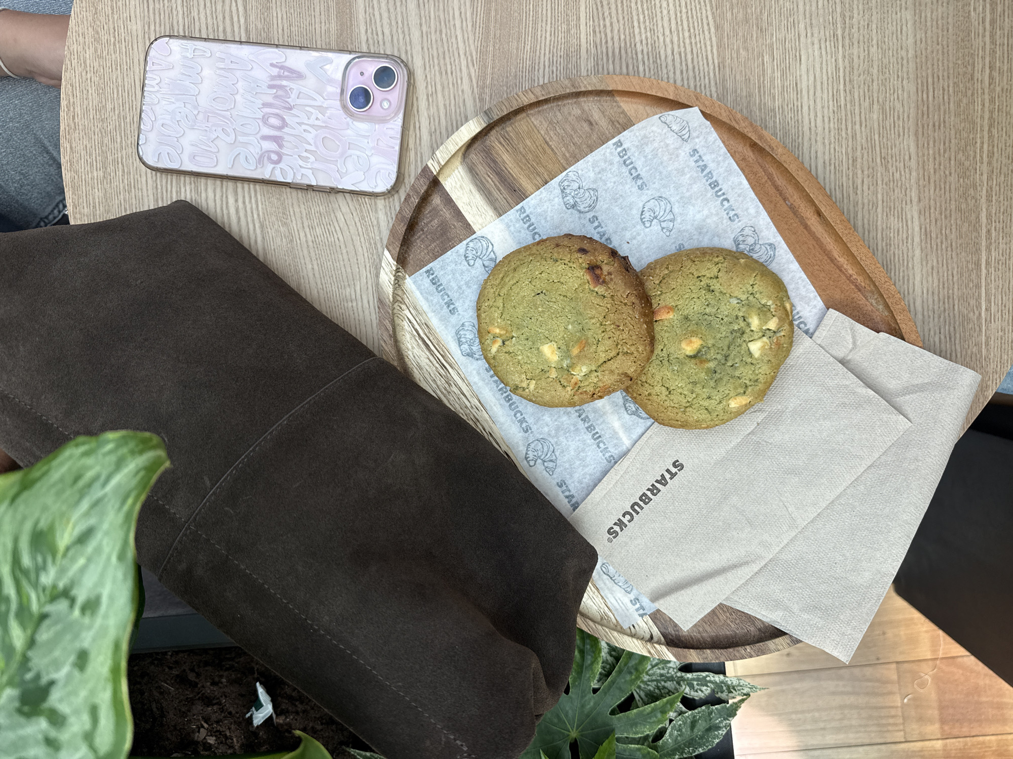 A high-angle shot shows a table setting in what appears to be a Starbucks in Matosinhos, Portugal. A mobile phone with a pink case displaying the word "Amore" sits to the upper left of the frame. A brown suede bag lies partially visible to the left. A wooden plate holds two green cookies, served on paper napkins with Starbucks logos and croissant illustrations. A stack of beige Starbucks napkins is also present on the plate. Green plants are situated below the table's surface, near the bag. The table itself has a light wood grain finish.