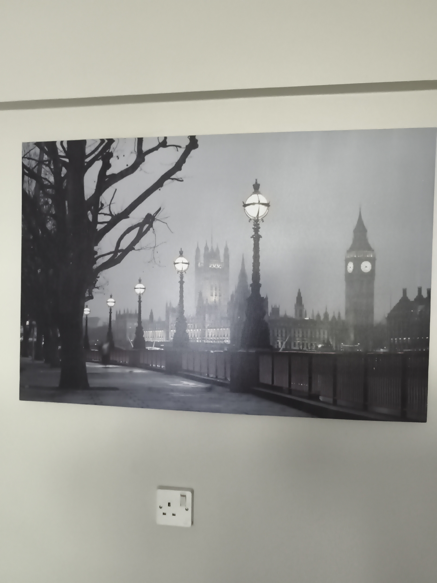 The media file presents a large, landscape-oriented canvas print of a monochrome cityscape, hanging on a plain, light-colored wall. Below the print, centered on the wall, is a white UK-style electrical wall socket with two outlets and a switch.
The canvas print depicts a classic view of London, specifically the Houses of Parliament and the Elizabeth Tower (Big Ben). The image is rendered in black and white or grayscale, imbuing it with a timeless, atmospheric quality.
In the foreground, on the left, a large tree with dark, bare branches dominates, with more trees lining what appears to be a walkway or embankment. Along the right side of this pathway, several ornate streetlights are visible, their globes emitting a soft, ethereal glow that illuminates the immediate surroundings and the bridge railing. The railing runs horizontally across the lower middle of the frame.
In the background, the intricate gothic architecture of the Houses of Parliament rises majestically. Many of its windows are lit, indicating an evening or nighttime scene. To the right, the iconic Big Ben clock tower stands tall, its illuminated clock faces clearly visible; the time appears to be approximately ten past ten. A soft mist or fog hangs over the cityscape, lending an almost painterly and somewhat melancholic atmosphere to the scene. The overall impression is one of quiet grandeur and historical significance. There are no discernible people or specific activities taking place. Architectural patterns on the Parliament building resemble large "XXX" markings. The location context indicates this image is from Huntingdonshire, United Kingdom, showing a popular British landmark.