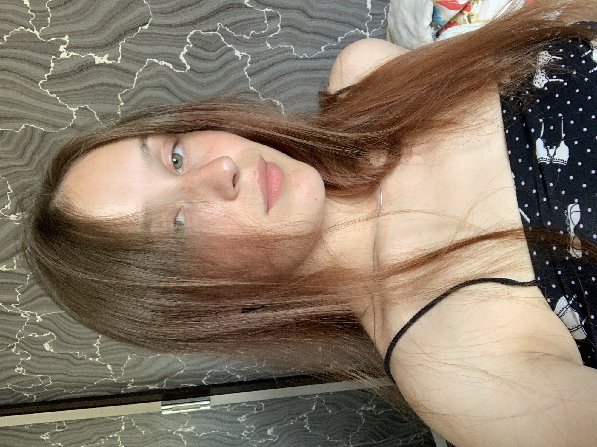 A young woman with light brown hair and freckles is shown from the chest up. She is wearing a black camisole with white polka dots and a pattern of what appears to be bras and bows. Her hair is long and extends past her shoulders, falling forward to partially obscure her face. Her eyes are open and looking towards the camera. She is positioned in front of a grey, textured wall with a marbled pattern. The lighting appears to be natural, casting soft shadows. The image was taken in Fokino, Russia.